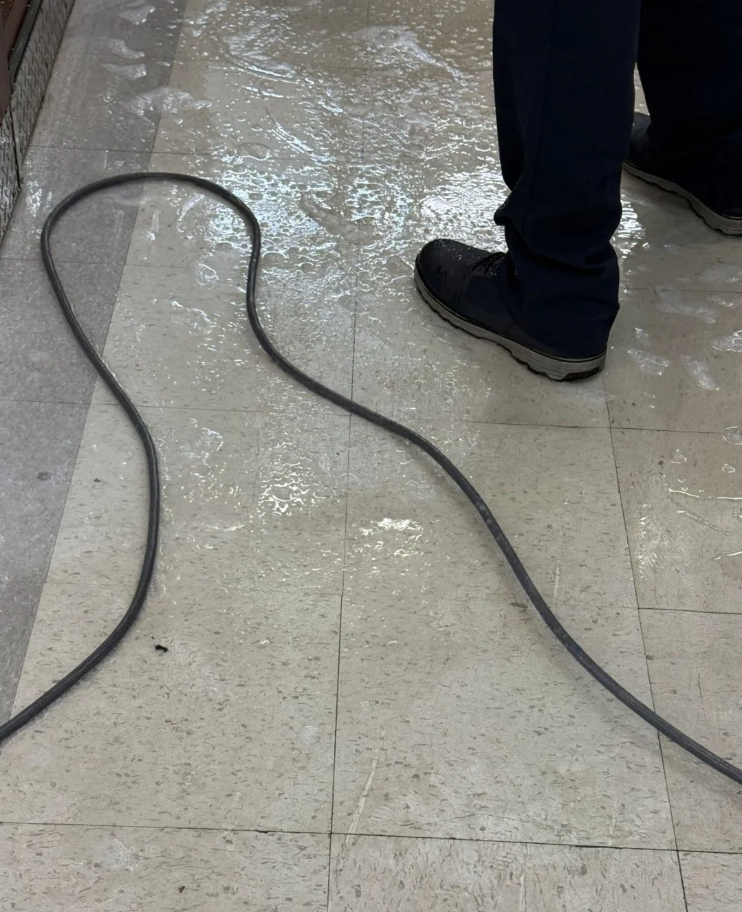 Person in black shoes and navy pants standing on wet beige tiled floor with a black cable on the floor, near a tiled wall.