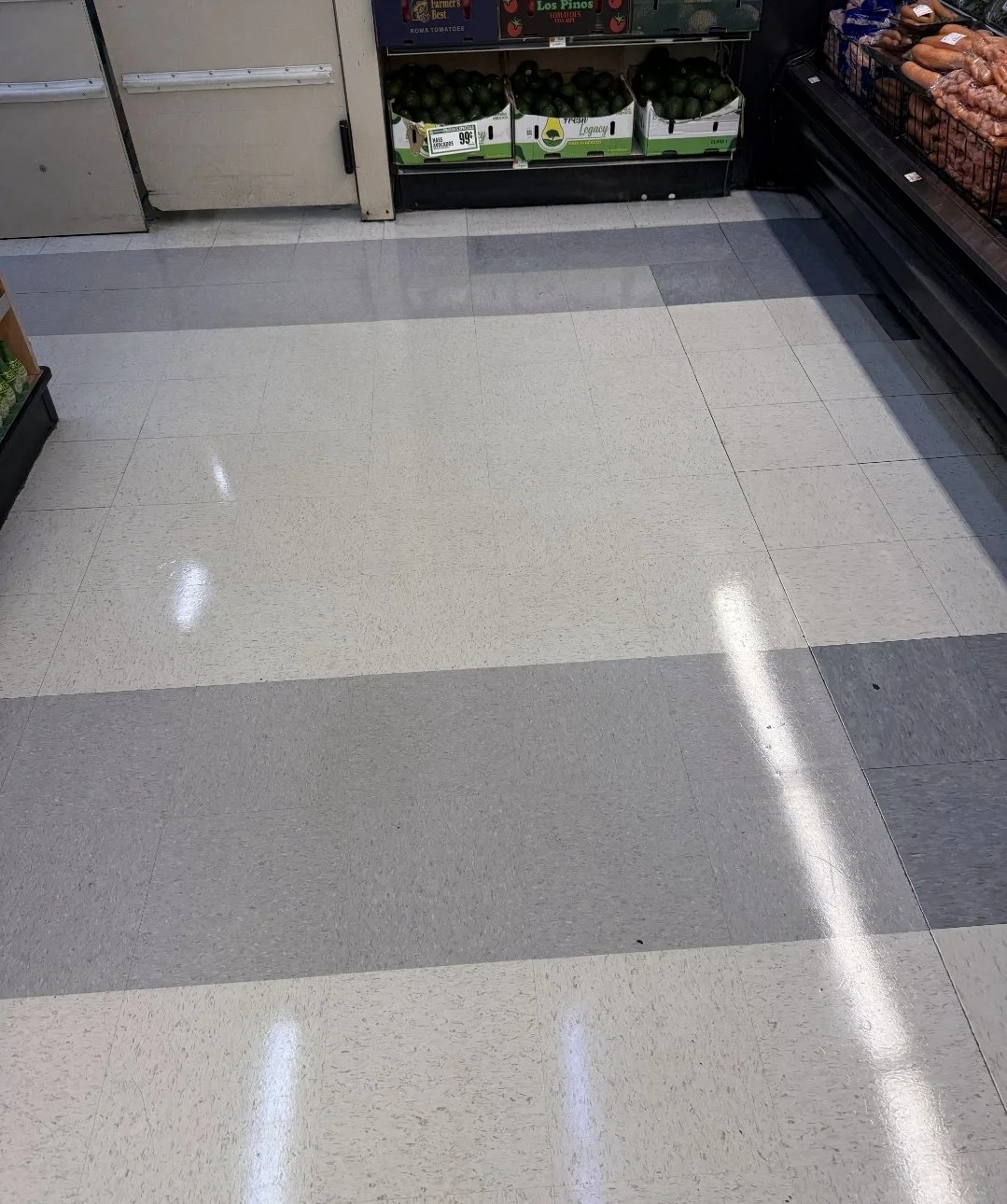 Empty grocery store aisle with a tiled floor in shades of beige and gray, and glass display cases with avocados and other produce on shelves in the background.