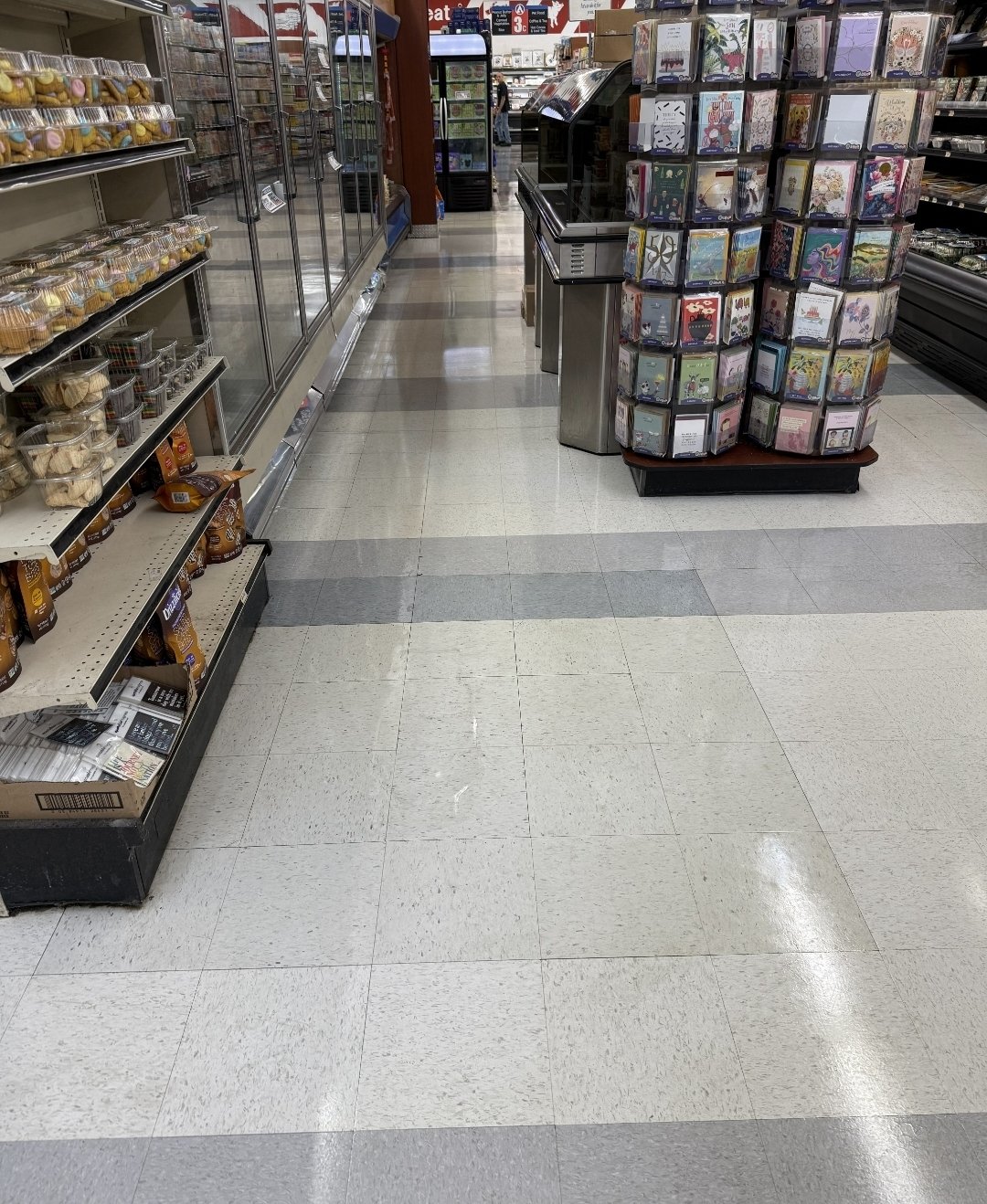 Empty grocery store aisle with shelves stocked with packaged food on the left and a rotating stand with greeting cards on the right.