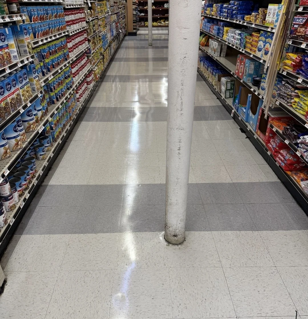 A grocery store aisle with a white pole in the middle, shelves stocked with various food products on both sides, and a shiny tiled floor.