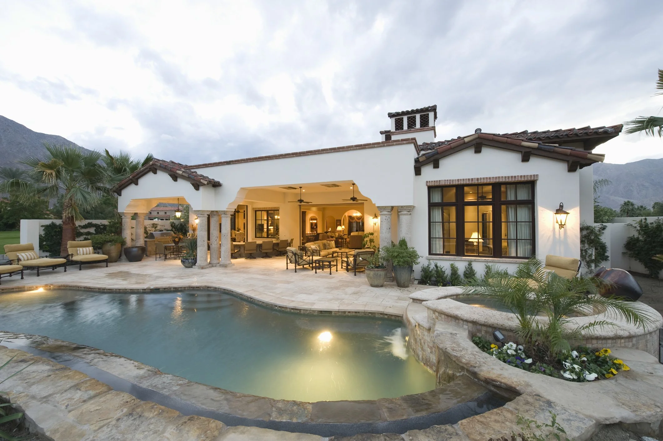 Exterior view of a house with a backyard featuring a swimming pool, outdoor seating area, and palm trees. The house has Spanish-style architecture with tile roof, large windows, and lantern-style lights.