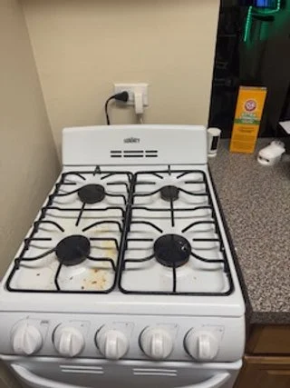 A white gas stove with four burners, some with black knobs and a few with burnt food spills, is positioned next to a kitchen countertop with various items on it, including a box, a white jar or container, and a coffee machine.