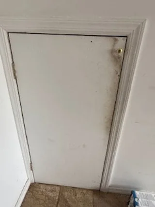 A small white door with dirt and marks, set in a white wall, with a brown tile floor in front.