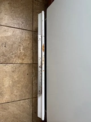 A white power strip with multiple outlets, plugged into a wall, placed vertically on a brown tiled floor.