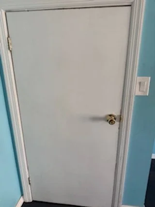A plain white door with a gold doorknob, set in a light blue wall with a white trim and a light switch nearby.
