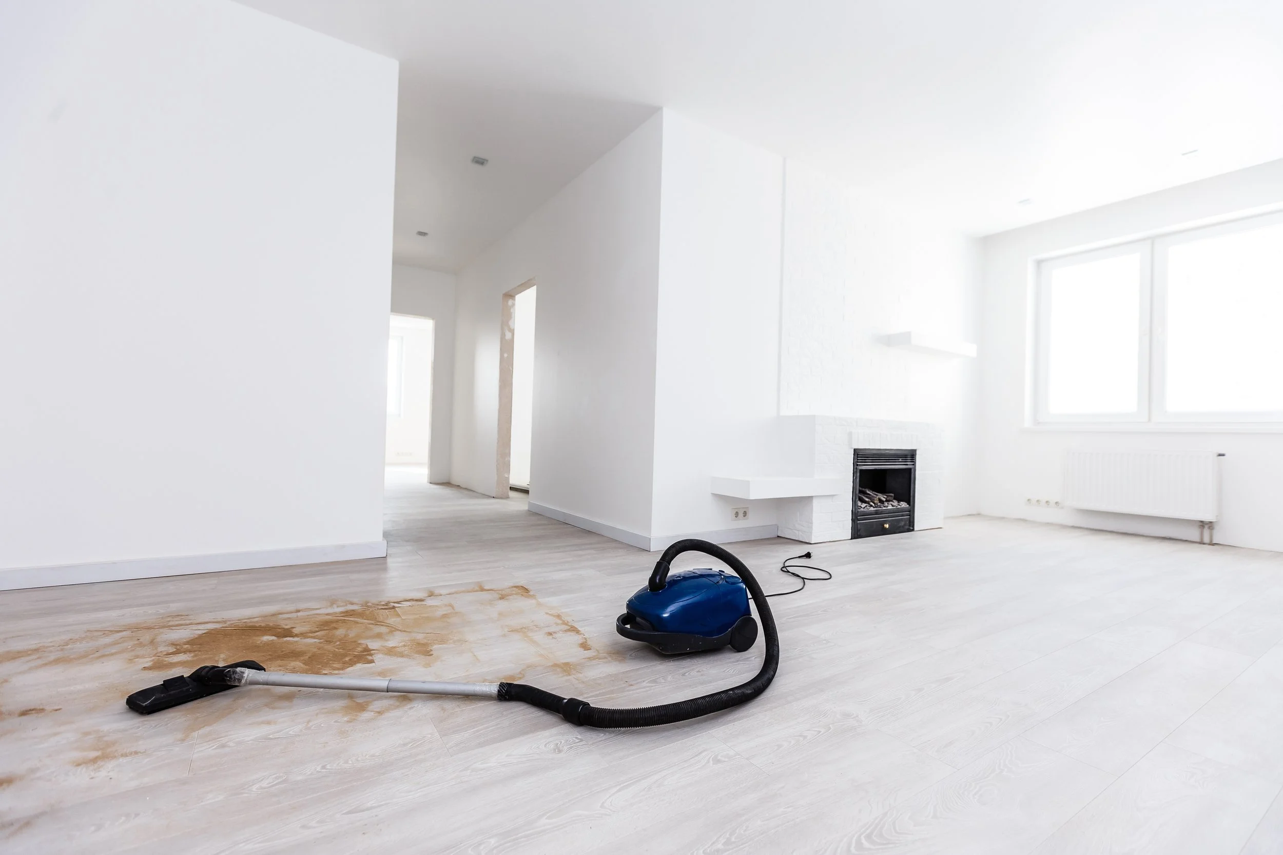 White living room with a fireplace, large windows, and hardwood floors with a broken section, a vacuum cleaner, and a blue canister on the floor.