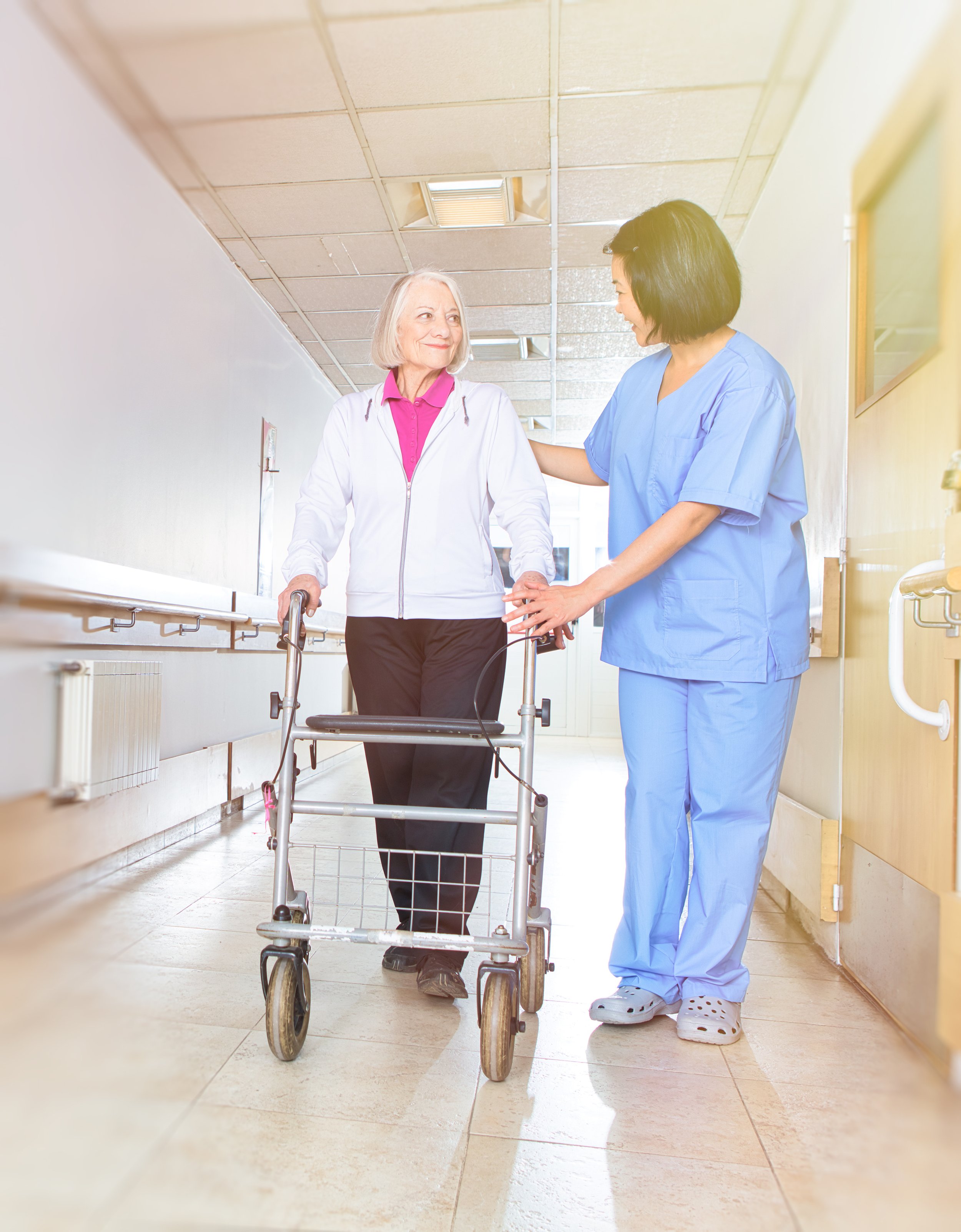 A healthcare worker in blue scrubs assisting an elderly woman with gray hair using a walker in a hospital corridor.