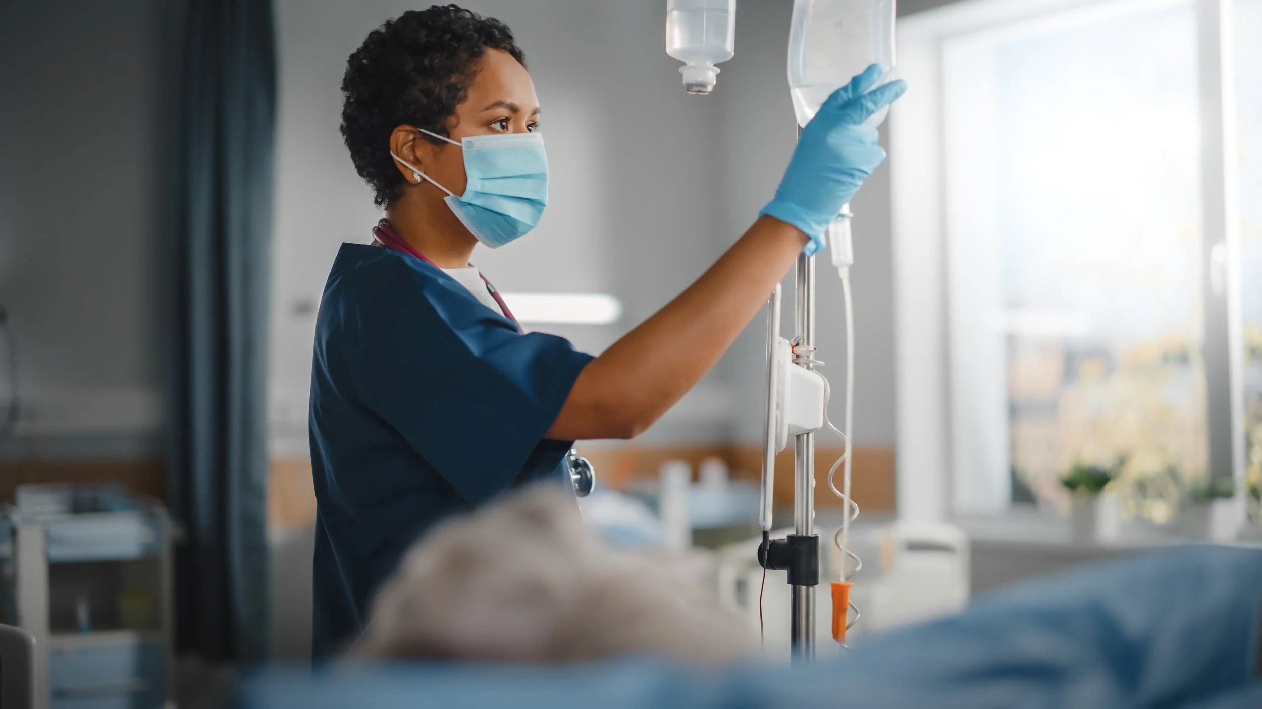 A healthcare worker wearing a face mask, gloves, and scrubs setting up or adjusting an IV drip in a medical facility with natural light.