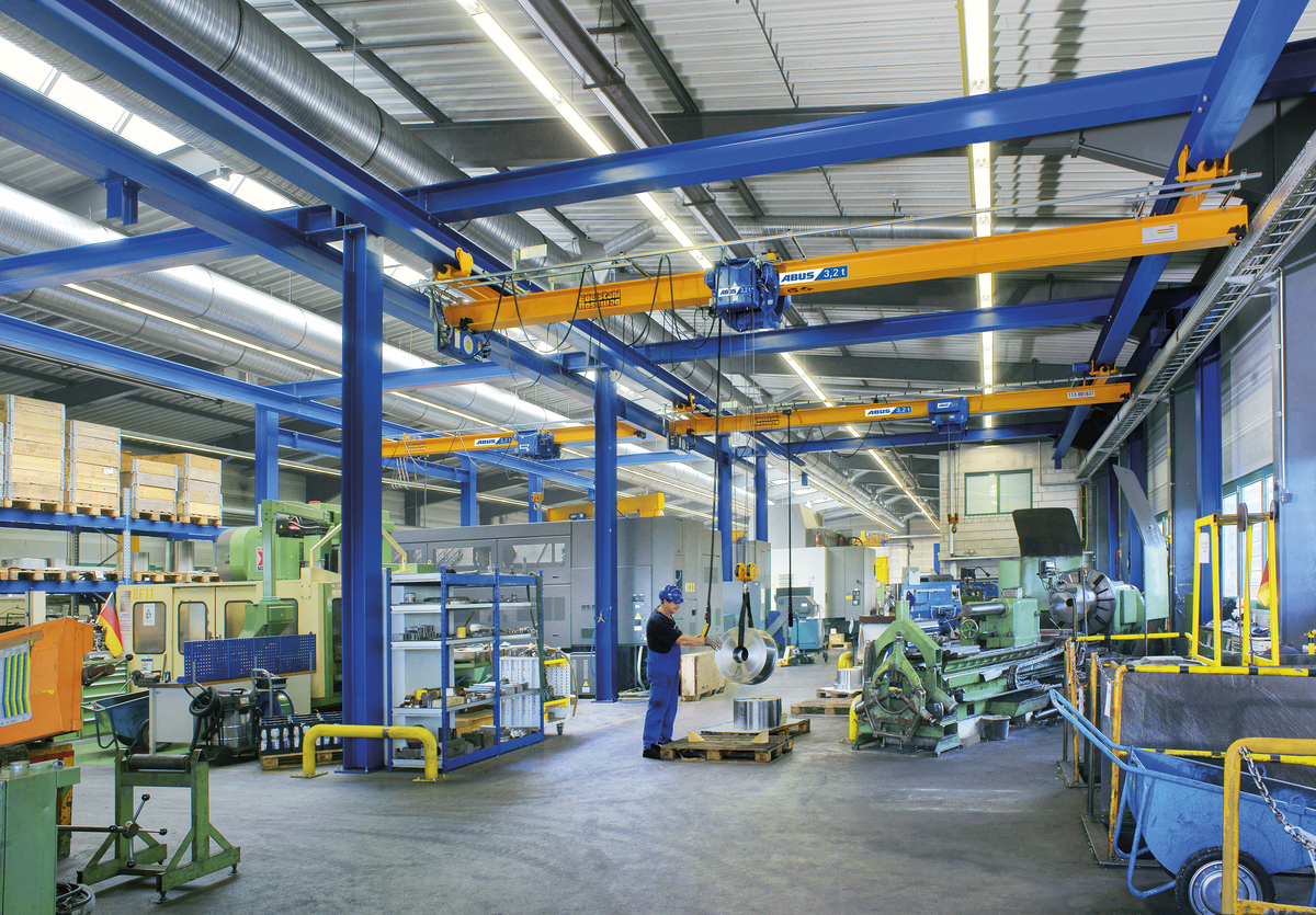 Interno di una fabbrica con macchinari industriali, un operatore al lavoro con un rotolo di metallo, gru gialle sospese, scaffalature con materiali e vari strumenti.