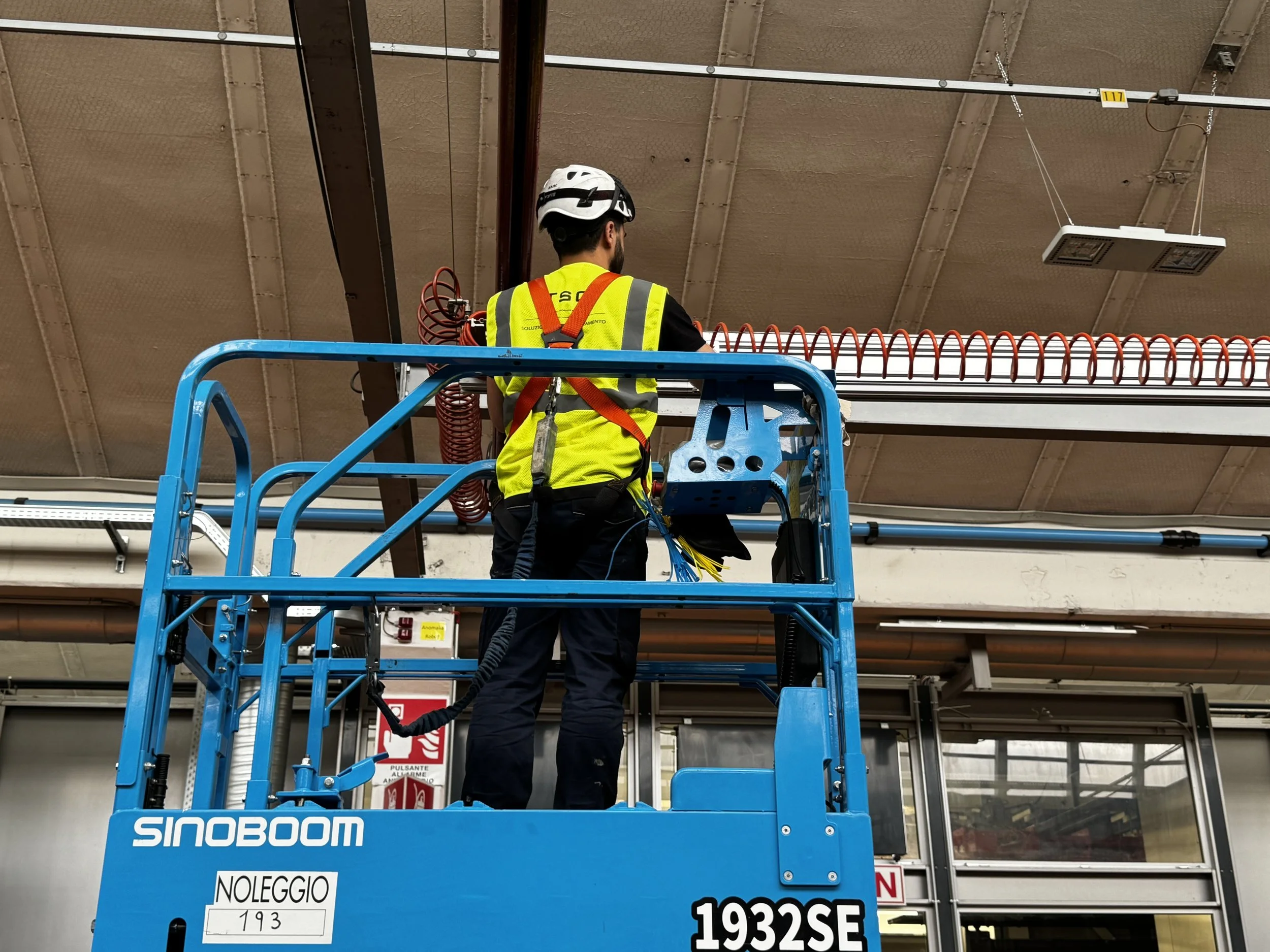 Un operatore sta lavorando sopra una piattaforma elevatrice blu con la scritta 'SINOBOOM', indossando un elmetto bianco e una giacca gialla con riflettori. Sta collegando o verificando cavi elettrici vicino al soffitto di un edificio commerciale o industriale.