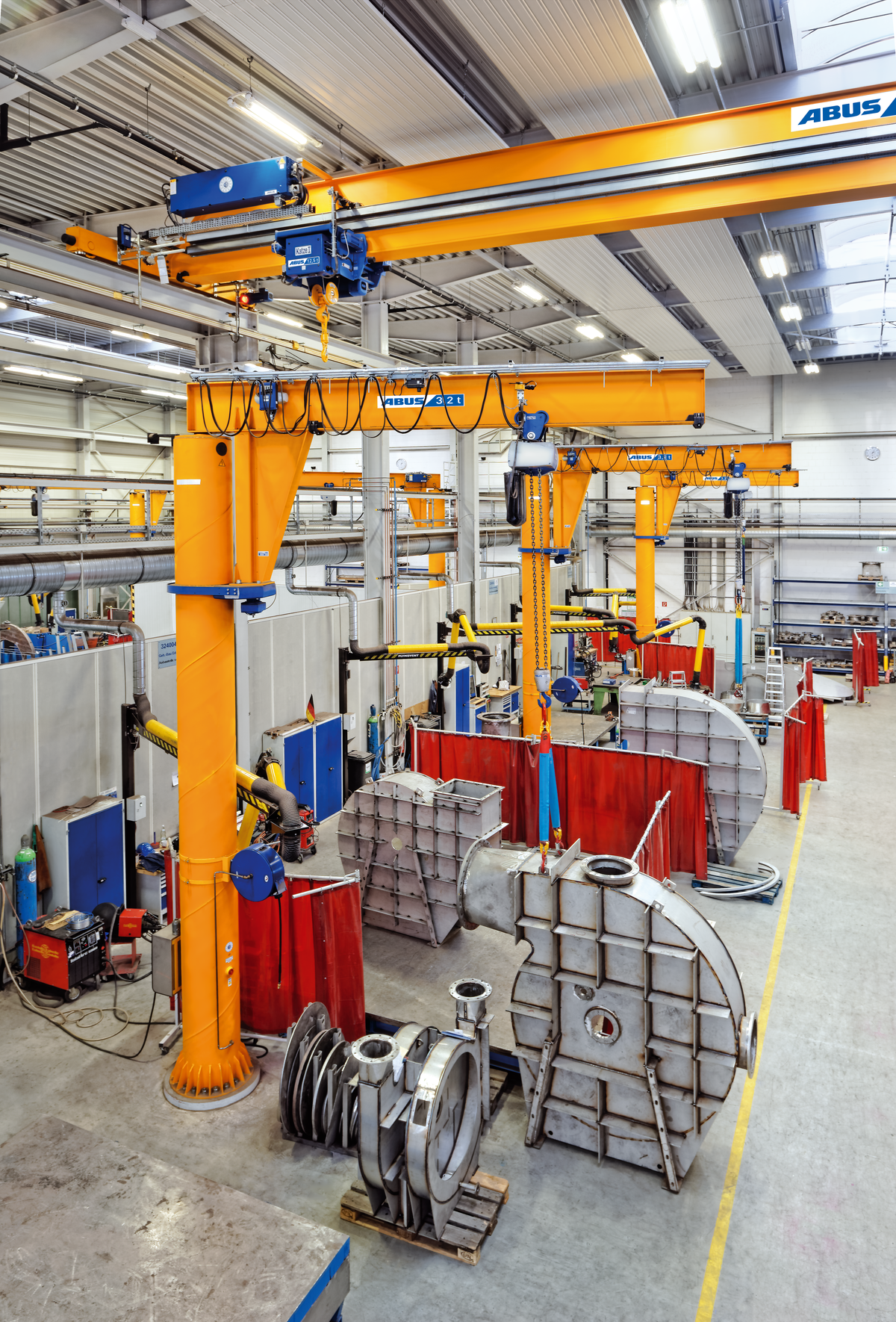 Interno di un'officina con grandi gru di colore arancione e diversi pezzi di grandi dimensioni, occhialati per la lavorazione industriale, e attrezzi vari visibili.