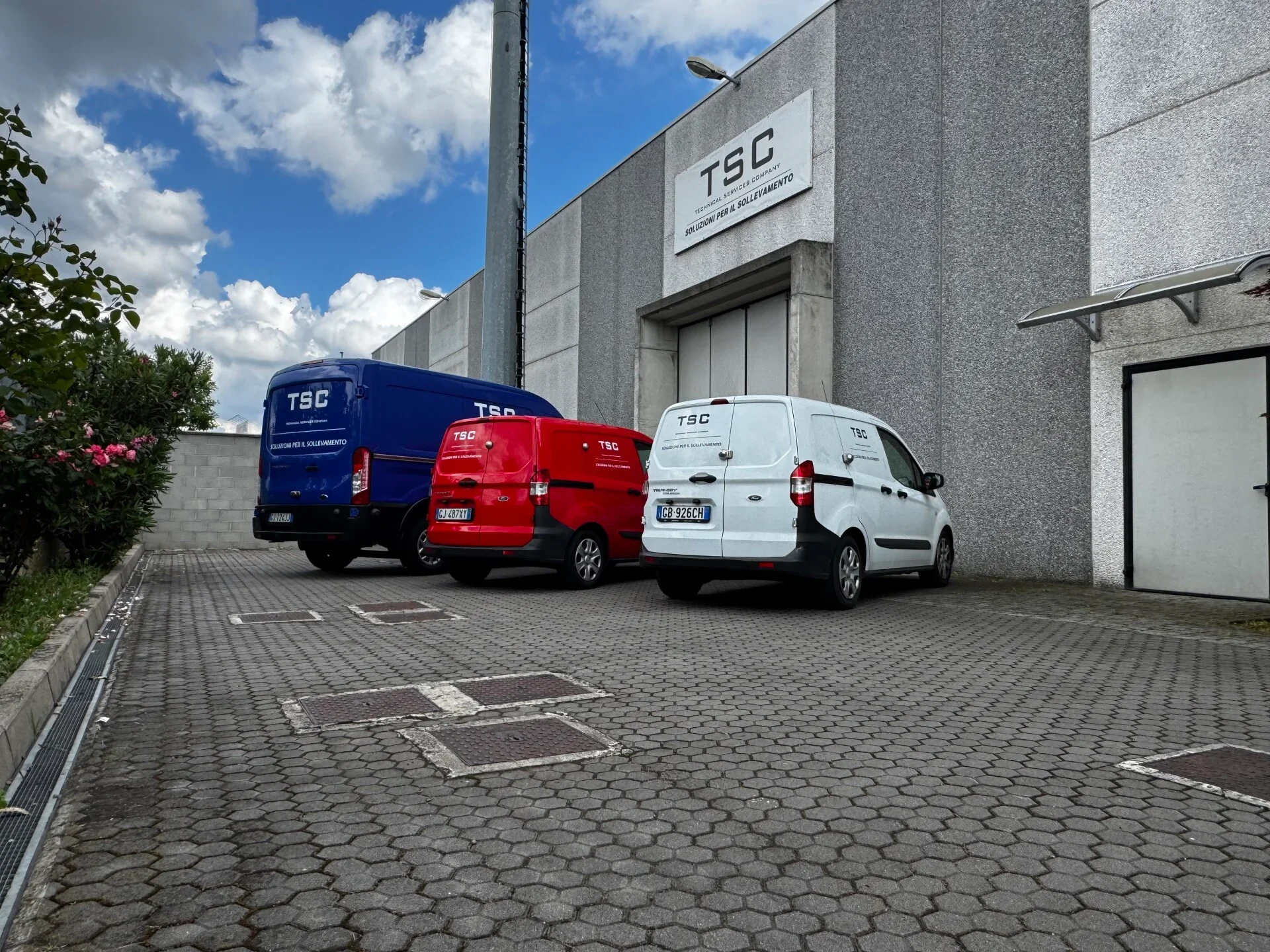 Tre veicoli commerciali bianco, rosso e blu parcheggiati davanti a un edificio con segnale 'TSC Soluzioni Per Il Sollevamento'. Cielo nuvoloso sopra. Pavimentato con mattonelle di cemento.