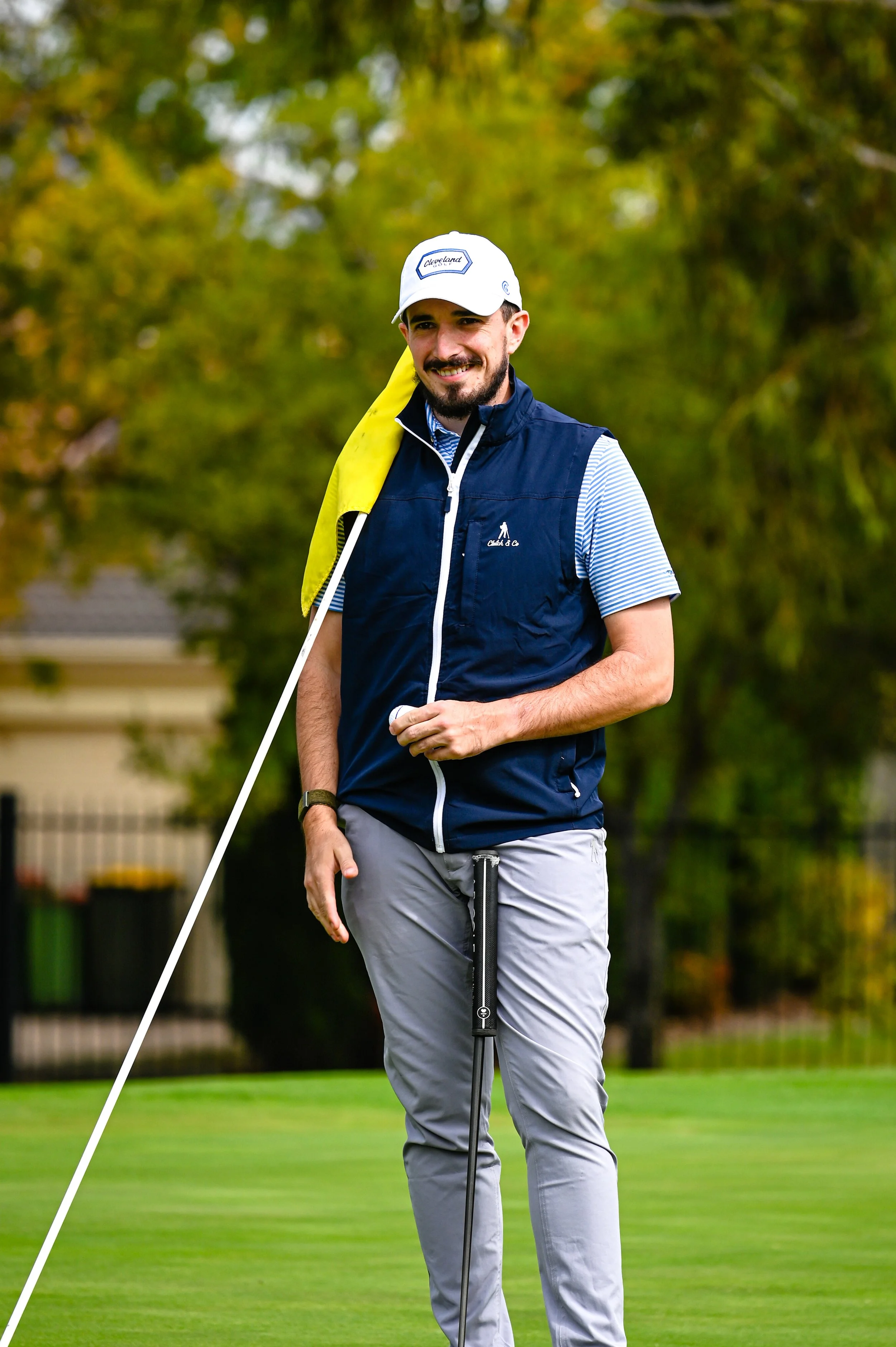 A man on a golf course holding a golf club and a yellow towel over his shoulder, wearing a white cap and a navy vest.