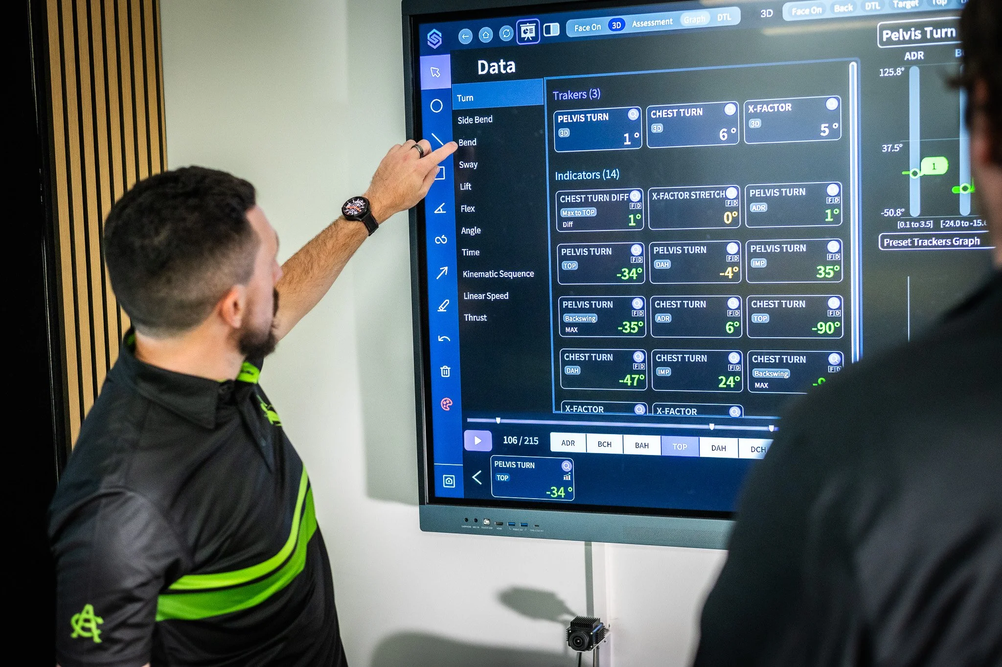 A person in a black shirt with green accents is pointing at a monitor displaying a data screen with various technical measurements and graphs, while another person watches.