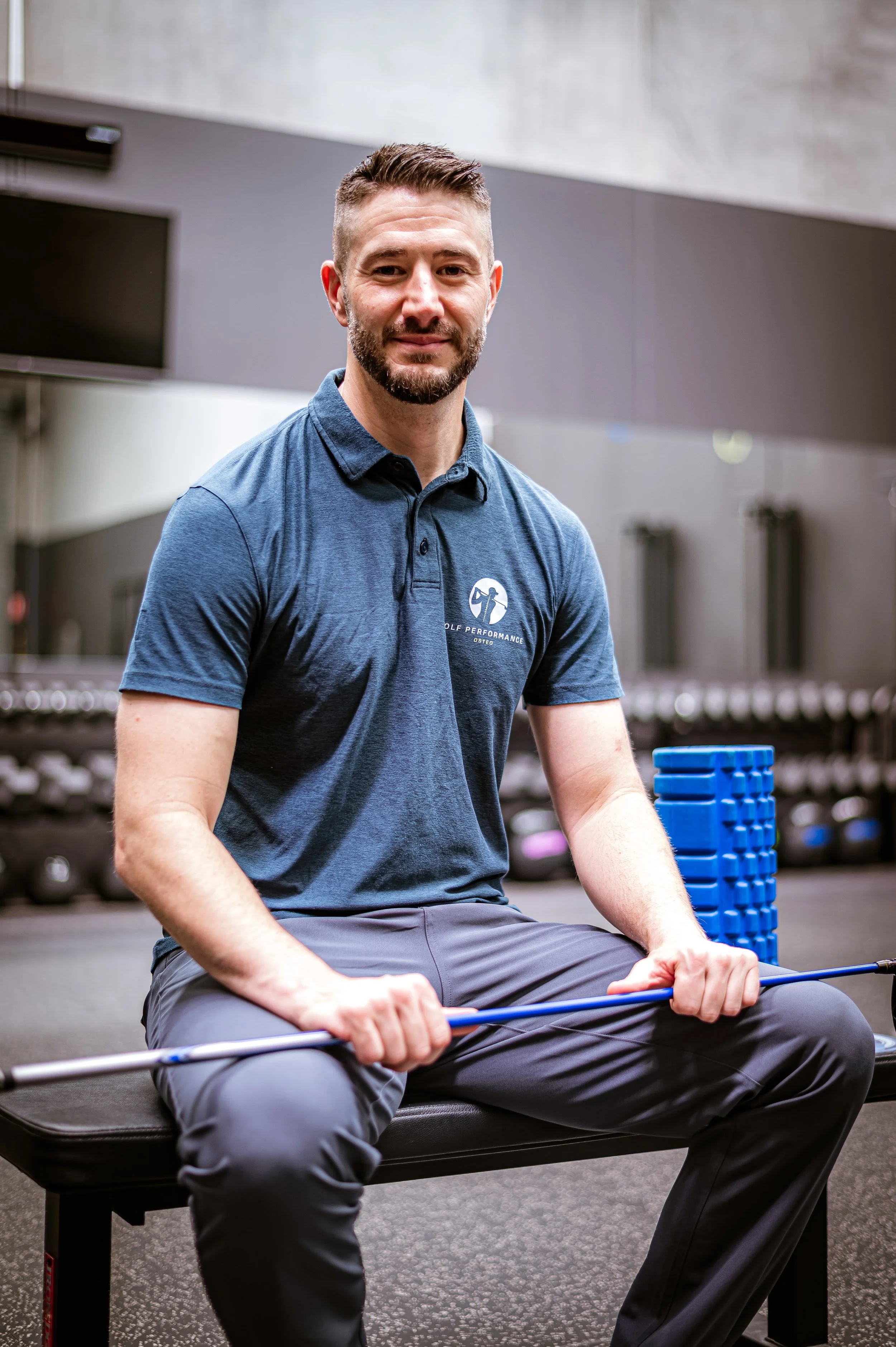 A man with a beard, wearing a blue polo shirt and gray pants, sitting on a black bench in a gym, holding a blue barbell with both hands, with a stack of blue weights nearby. The background includes gym equipment and a gray wall.