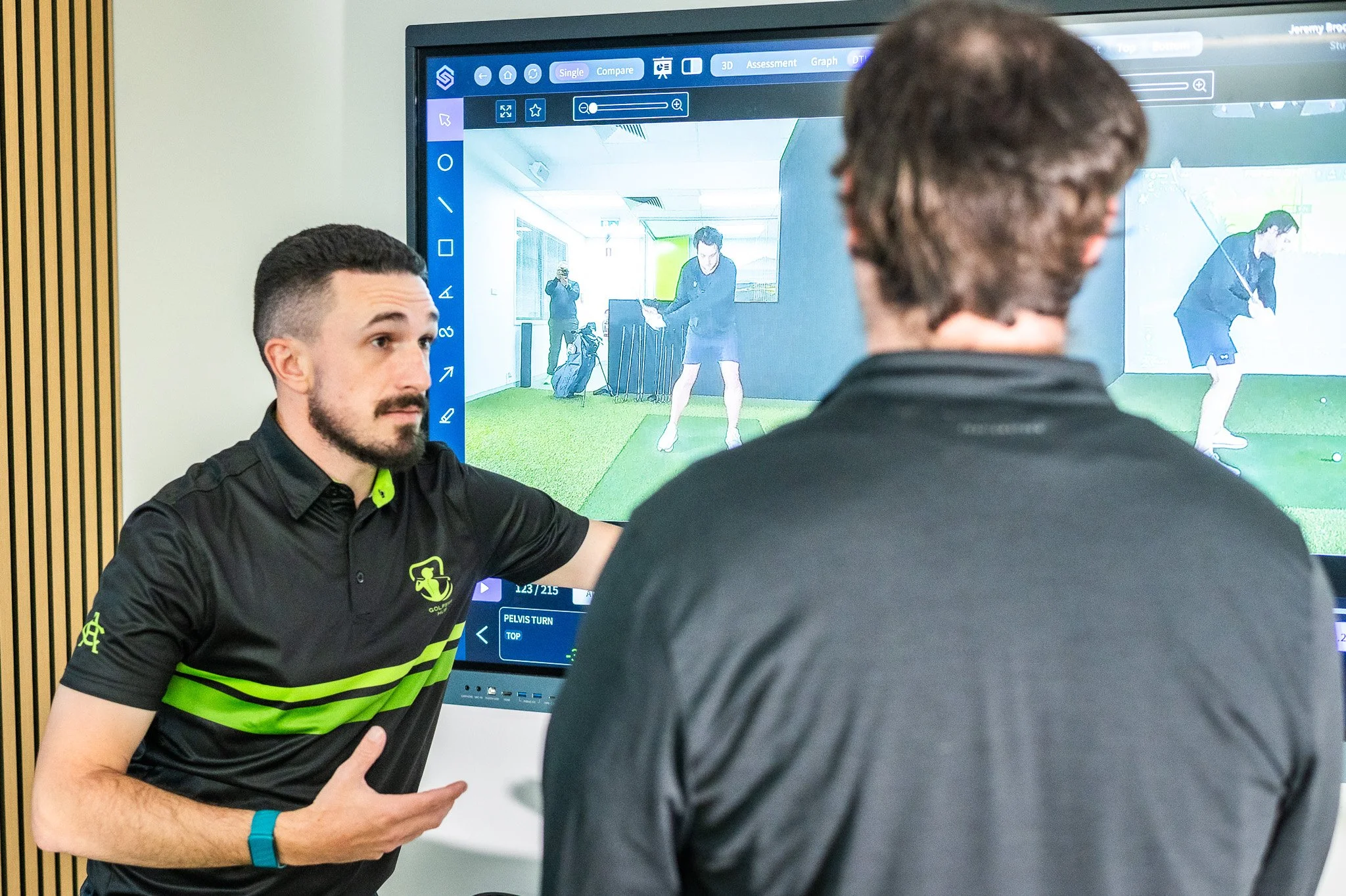 Two men in black and green golf attire having a discussion in front of a large display screen showing golf swing analysis images.