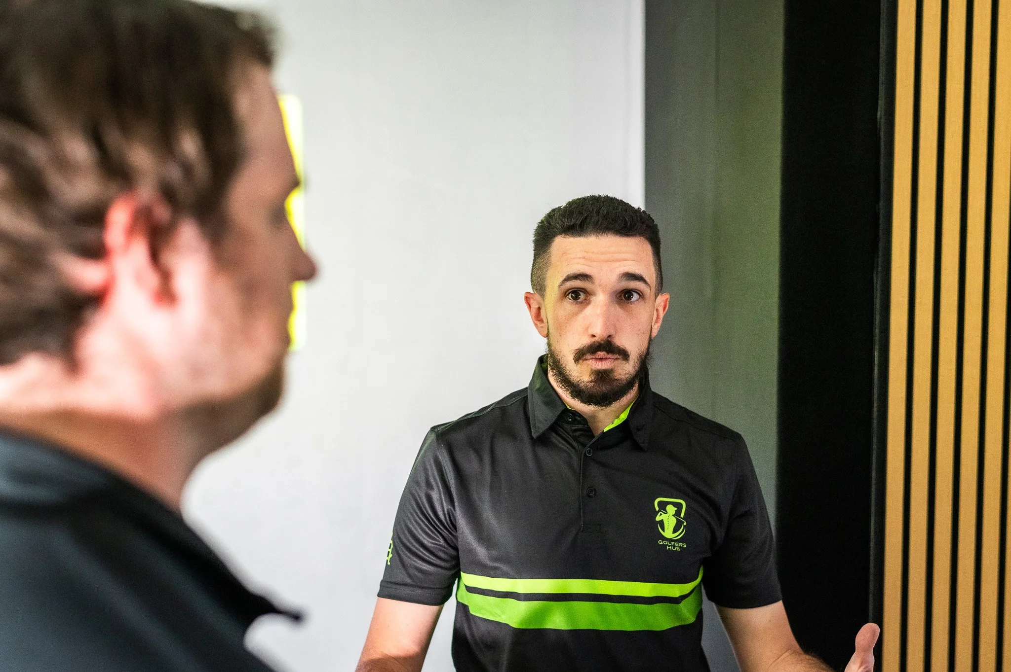 A man with dark hair, beard, and wearing a black shirt with neon green accents and logo, standing indoors near a vertical black and yellow striped wall, looking at a person in front of him with blurred face.
