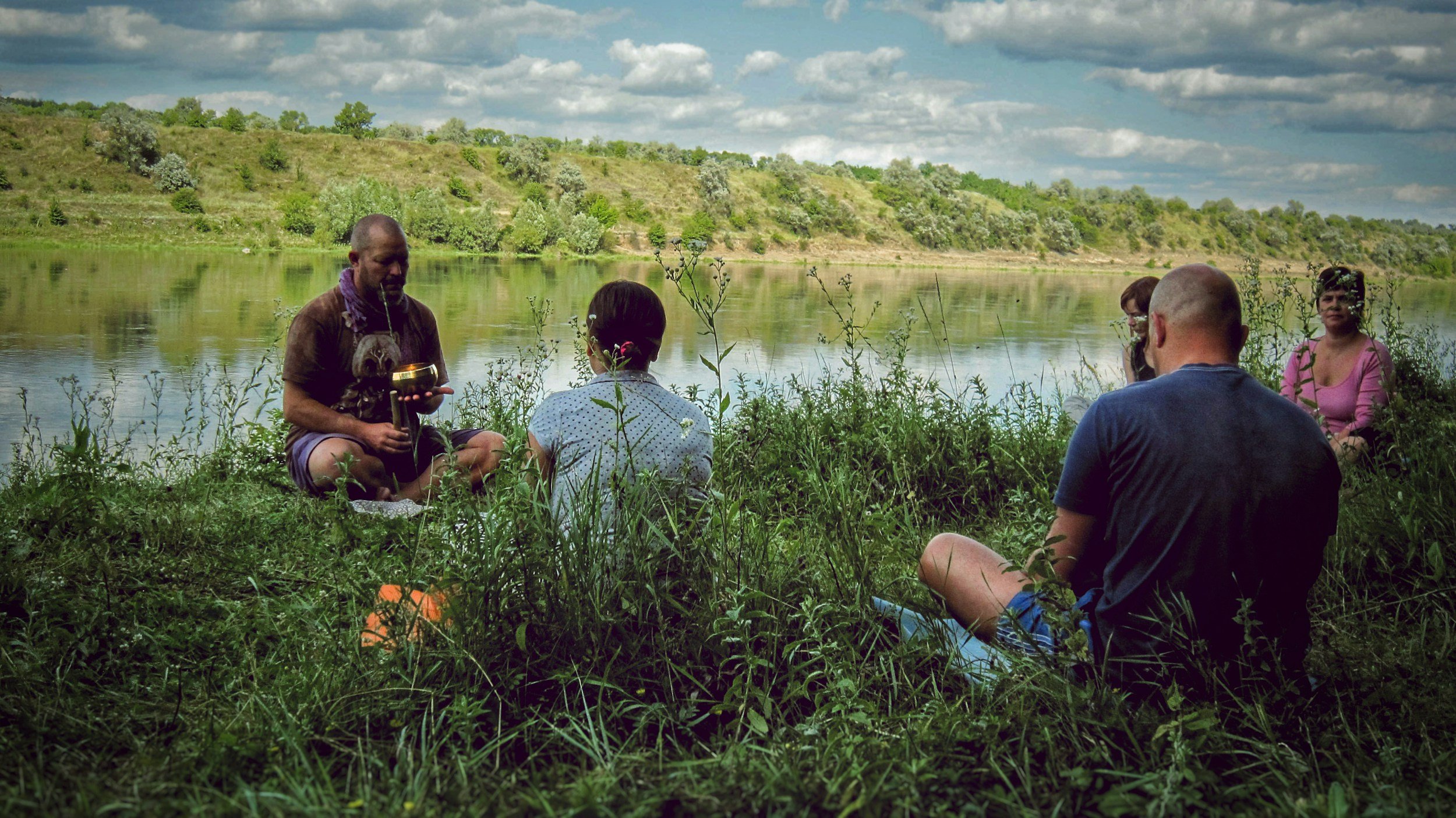 Group of five people sitting by a river in a grassy area, with a man holding an object, possibly participating in a discussion or meditation, and hills with trees in the background under a cloudy sky.