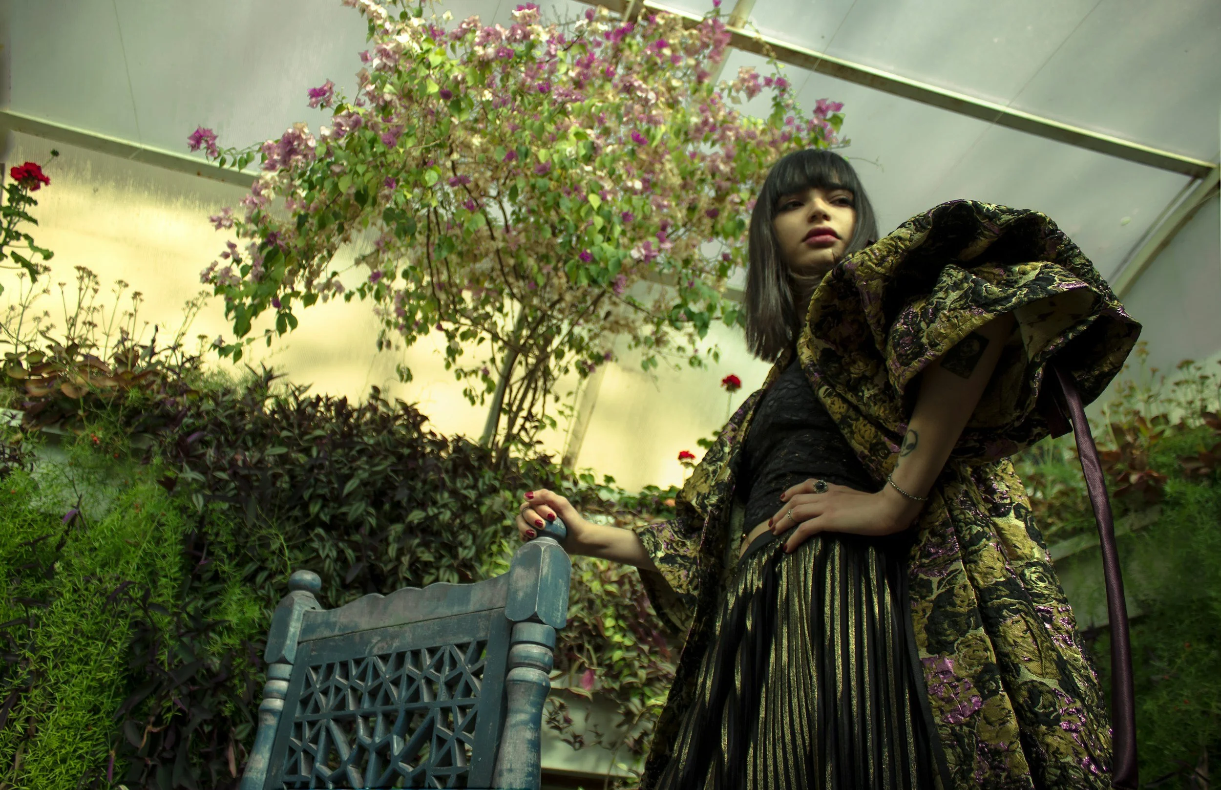 A woman in a floral jacket bending over in an indoor garden with various plants, including blooming bougainvillea, and a blue bench in the foreground.