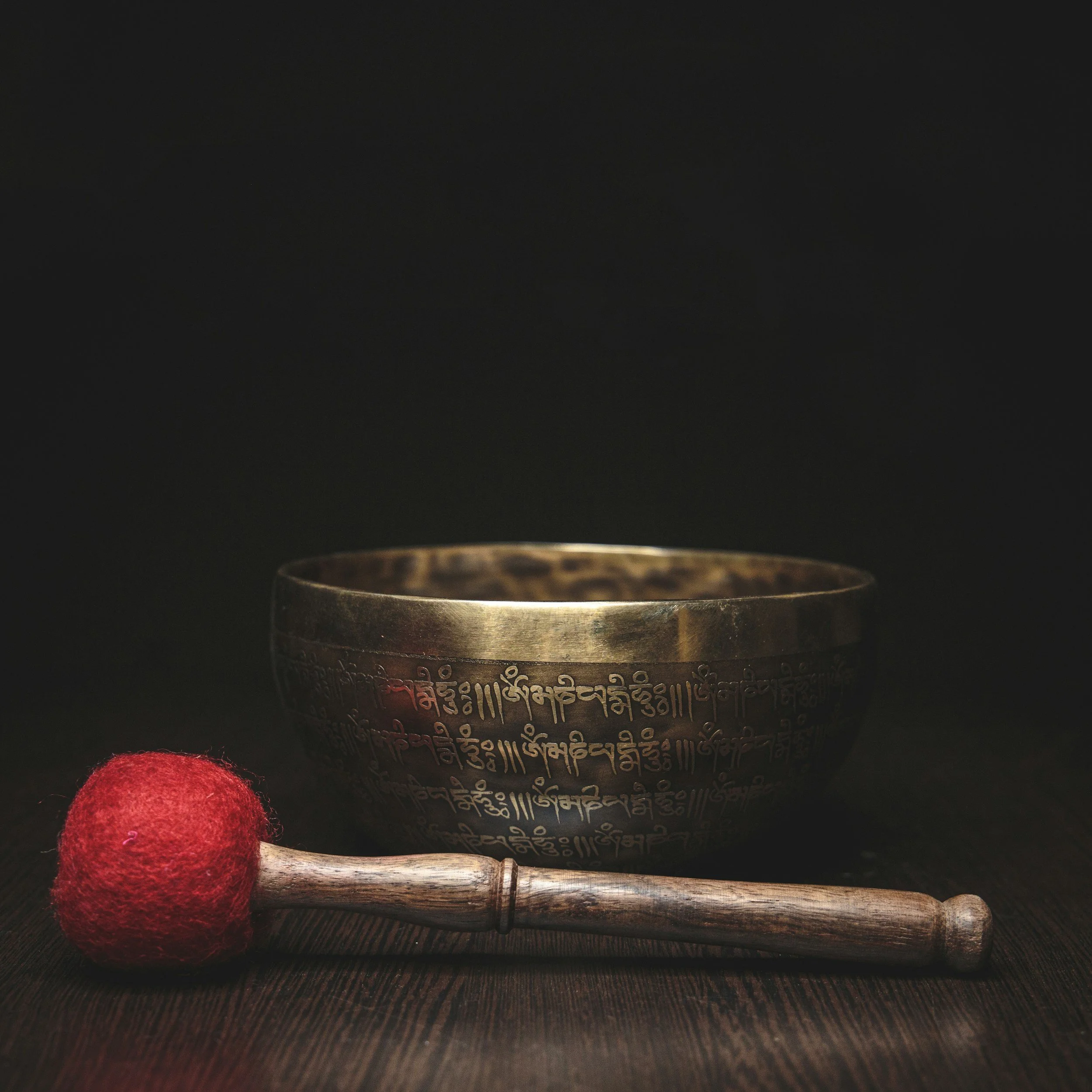 A Tibetan prayer wheel with Tibetan script engraved on its surface, placed on a wooden stand, with a red felt ball and a wooden stick in front of it, on a dark background.
