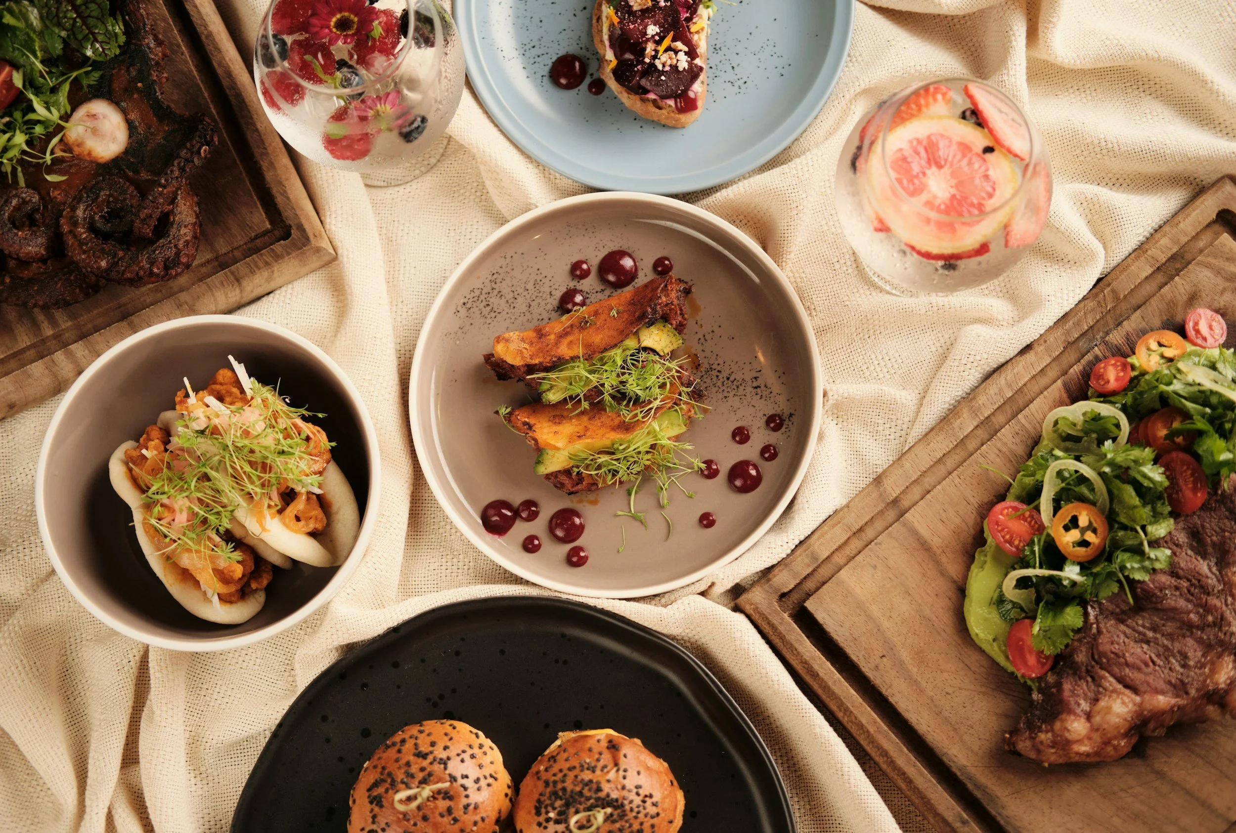 A variety of foods on a table including sliders with sesame seed buns, a salad with cherry tomatoes on toast, a plate of roasted chicken with greens, a slice of berry tart on a blue plate, two glasses of water with fruit slices, and a wooden tray of pretzels.