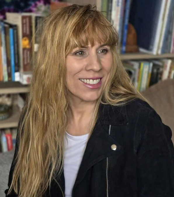 A woman with long blonde hair and a black jacket sitting in front of a bookshelf filled with books.