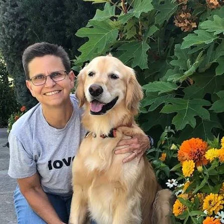 A smiling woman with short dark hair and glasses, wearing a gray T-shirt, sitting next to a large Golden Retriever in front of green leafy plants and colorful flowers.