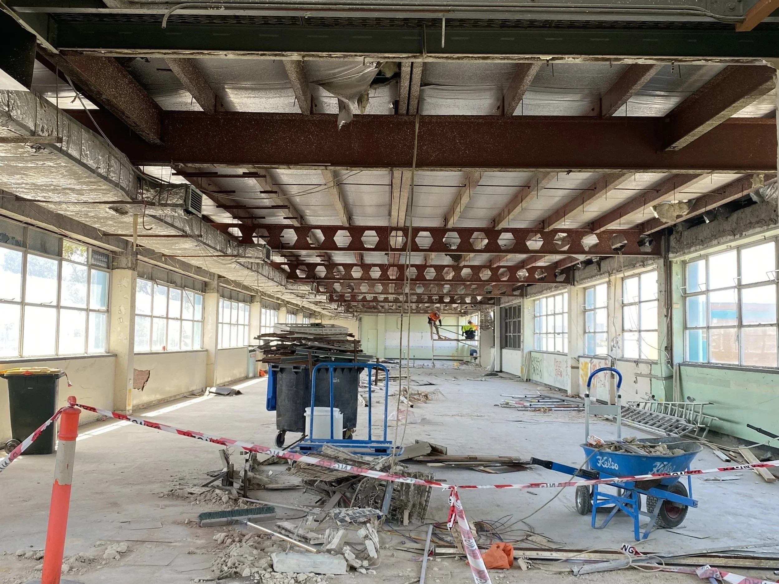 Interior of a one level of the BRUDI project in Brunswick under construction with exposed ceiling beams, construction workers, tools, and equipment scattered across the space, and safety barriers and tape.