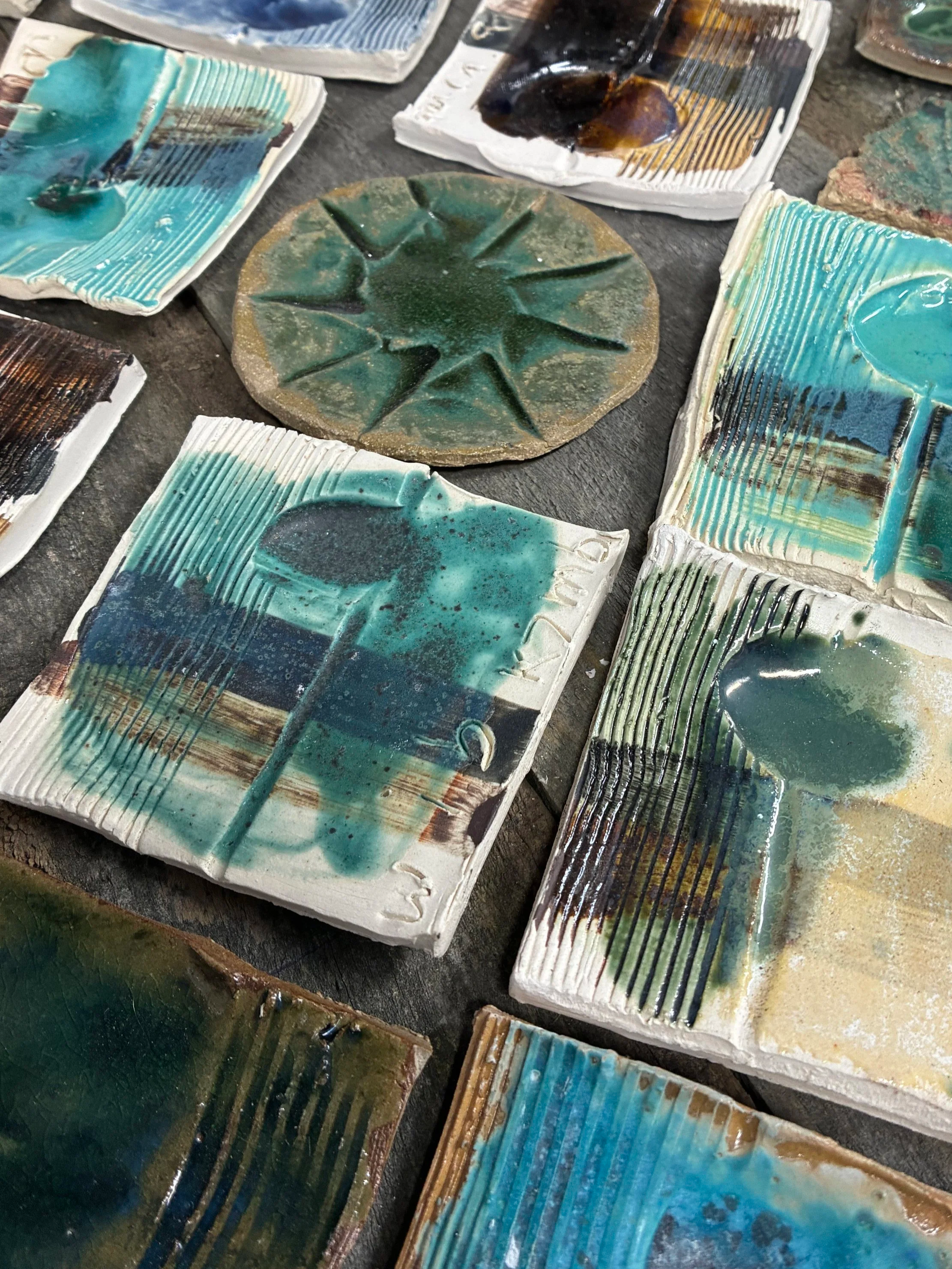 Close-up of glazed ceramic tiles with various colors and textures, arranged on a surface, some with linear patterns and others with abstract designs.