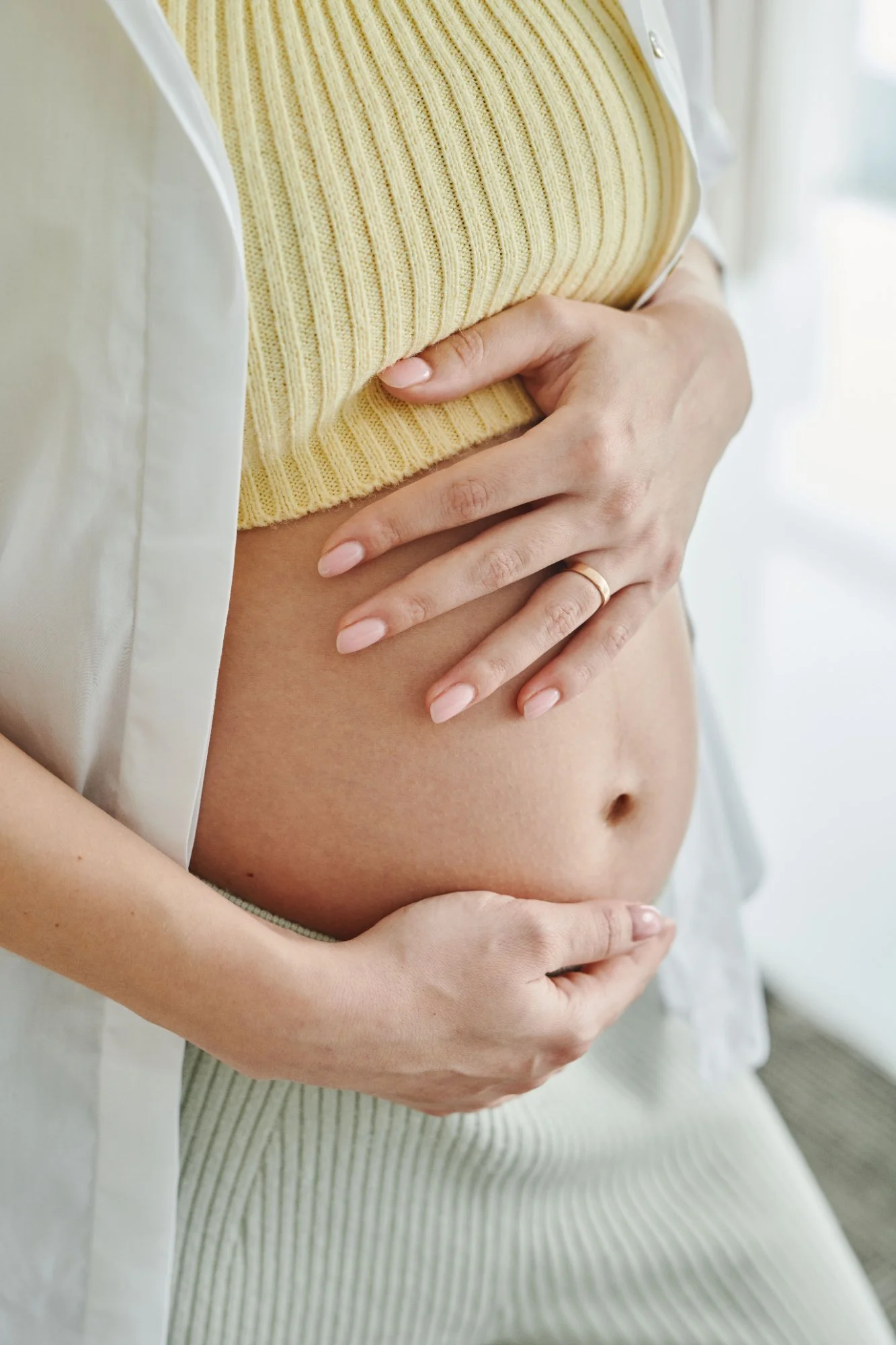 Close-up of a pregnant woman holding her bare belly with both hands, wearing a yellow knitted top and a light-colored cardigan.