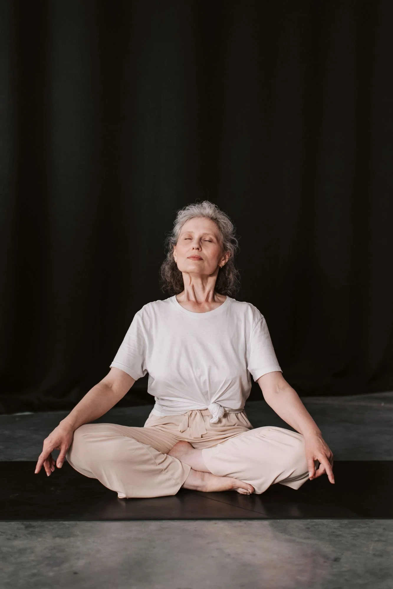 An older woman with gray hair practicing yoga or meditation in a seated cross-legged pose against a black background, eyes closed, with a peaceful expression.