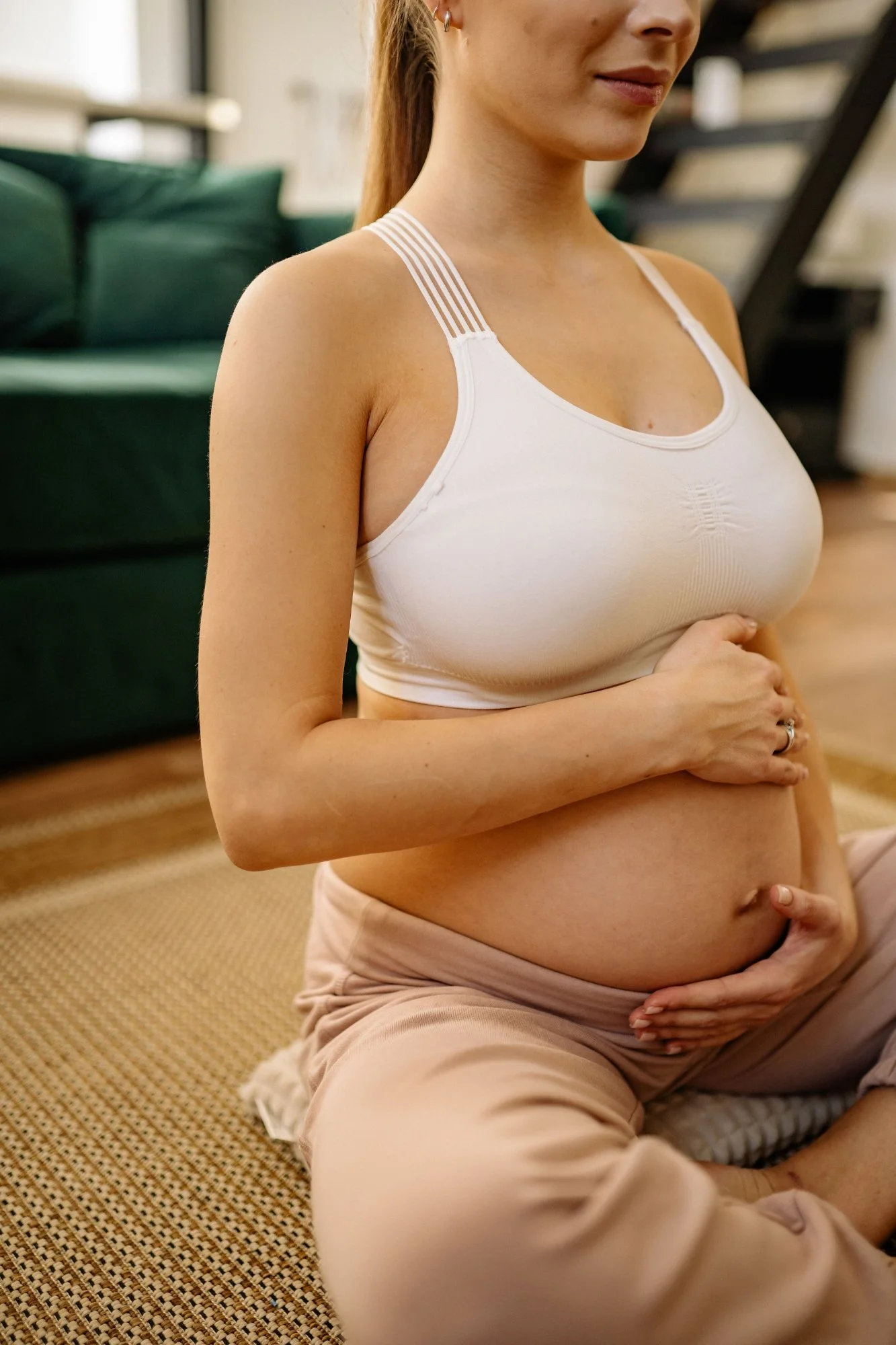 A woman practicing meditation indoors, seated on a rug with her hands resting on her abdomen, wearing a white sports bra and beige pants.
