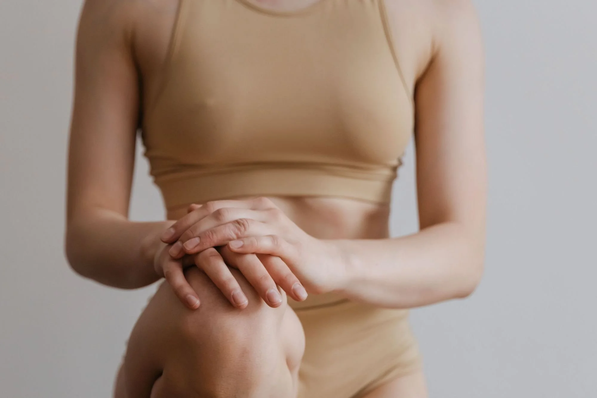 A person in minimal clothing with their hands clasped in front of their abdomen, standing against a plain neutral background.