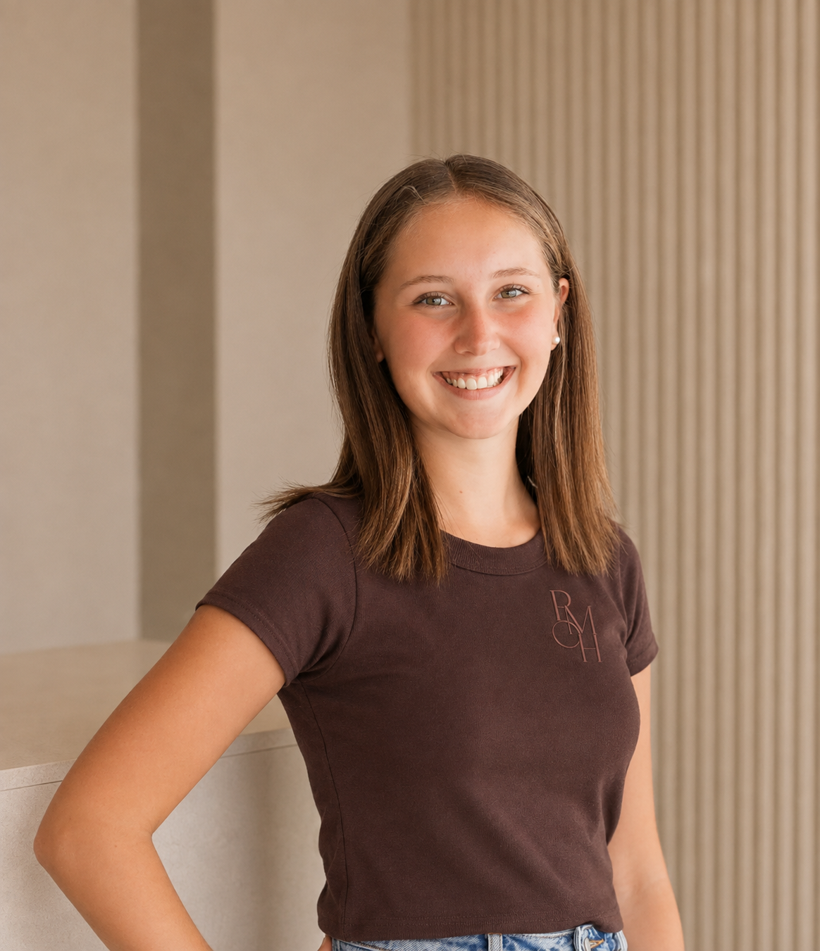 Smiling young woman with brown hair and blue eyes, wearing a dark brown t-shirt and standing in front of a beige wall with vertical stripes.