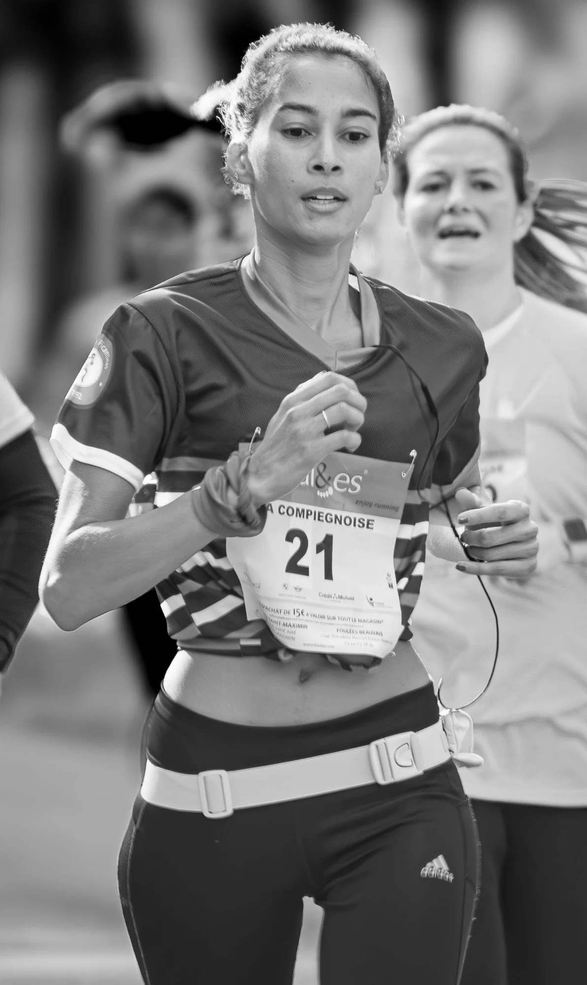 Black and white photo of a female marathon runner with race number 21, running outdoors with fitness clothing. She is wearing a sports bra, shorts, and a waist belt, with other runners visible in the background.