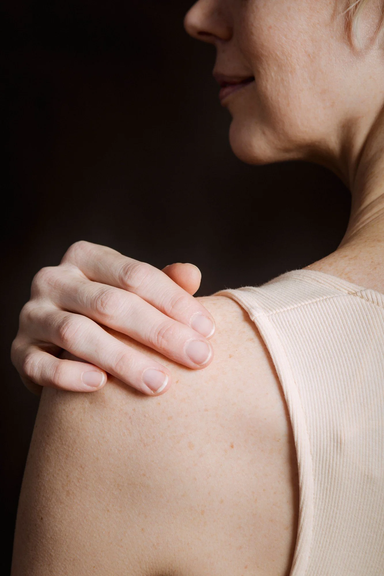 Close-up of a woman with fair skin and short blond hair, scratching her shoulder, wearing a beige sleeveless top, with a dark background.