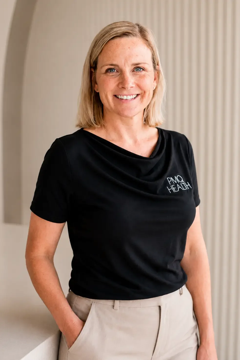 A woman with shoulder-length blonde hair, blue eyes, and light skin is smiling and standing indoors. She is wearing a black T-shirt with white text that reads 'PMQ HEALTH' and beige pants with one hand in her pocket.