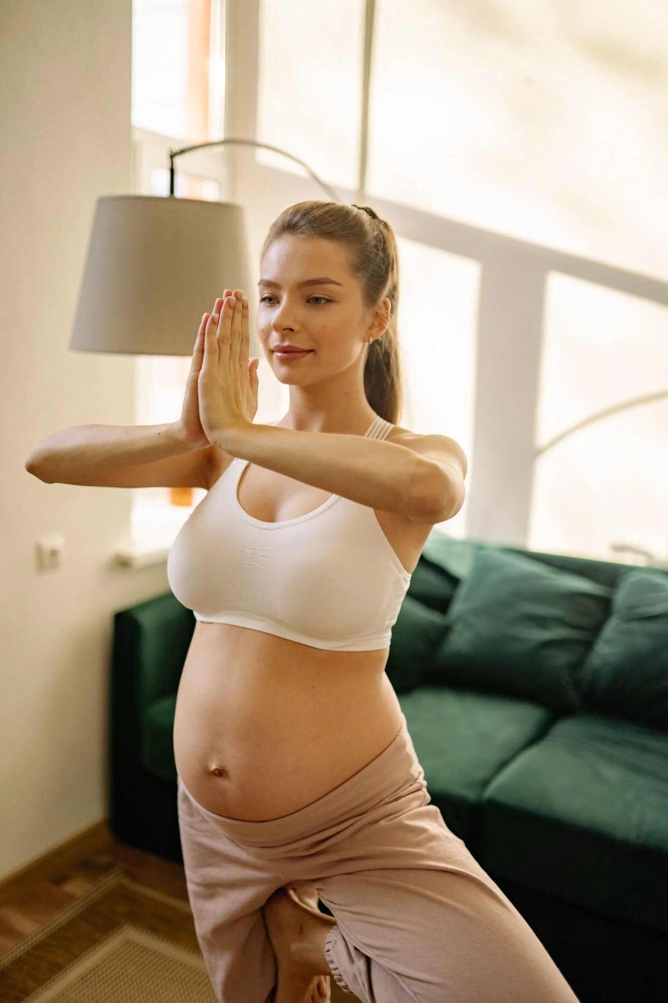 A woman practicing yoga indoors, standing in a pose with her hands together in a prayer position at her face, wearing a beige sports bra and beige pants, in front of a green sofa with pillows, with a lamp and window in the background.