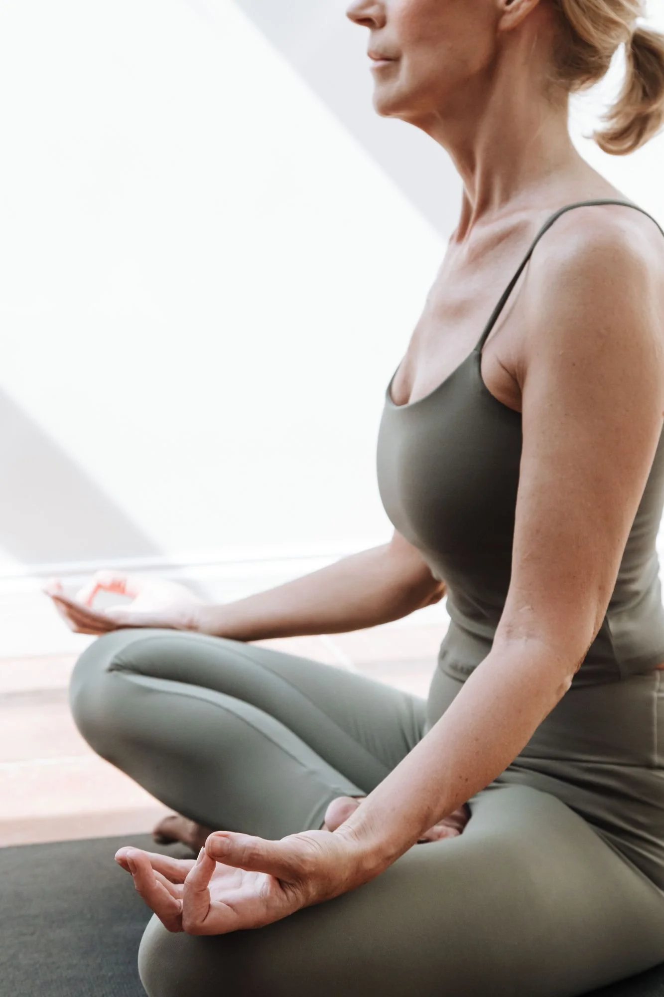 A woman practicing yoga in a seated meditation pose, with her legs crossed and palms resting on her knees, in a bright, minimalist room.