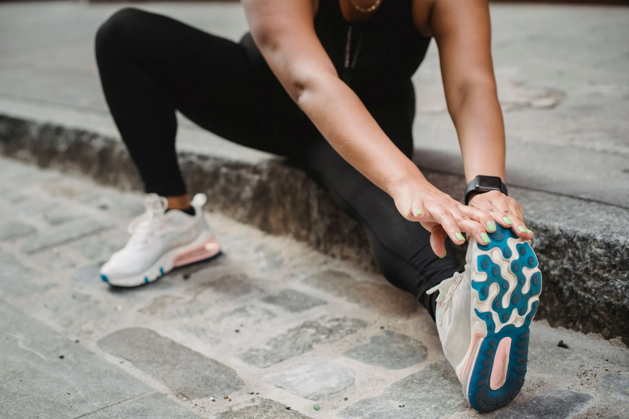 A woman in black leggings and white athletic shoes is bending over to stretch or adjust her sneaker on a cobblestone street. She is wearing a fitness tracker or smartwatch on her wrist.
