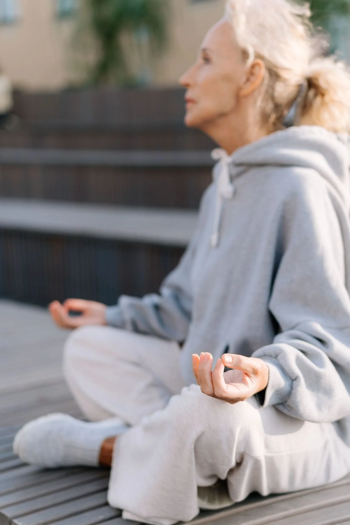 A woman practicing meditation outdoors, sitting cross-legged on a wooden surface with her eyes closed and hands resting on her knees in a peaceful pose.