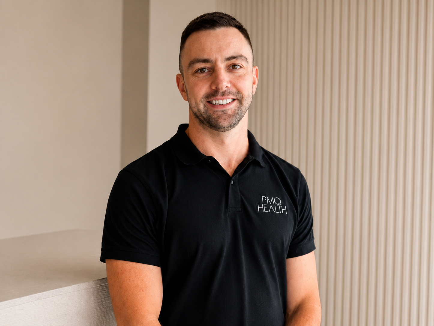 A man with short dark hair and a beard, smiling, wearing a black polo shirt with 'PMQ Health' logo, standing indoors near a wall with vertical wooden paneling.