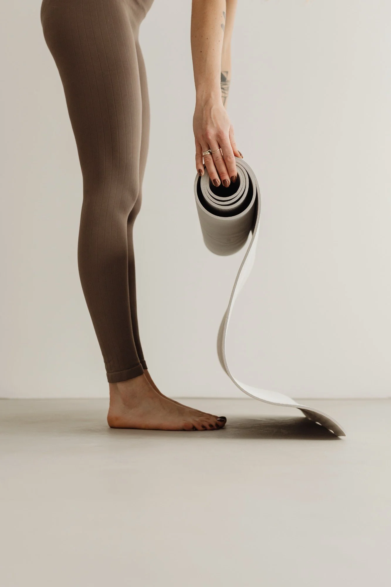 A person bending forward and holding a rolled yoga mat, standing barefoot on a beige floor against a plain white wall.