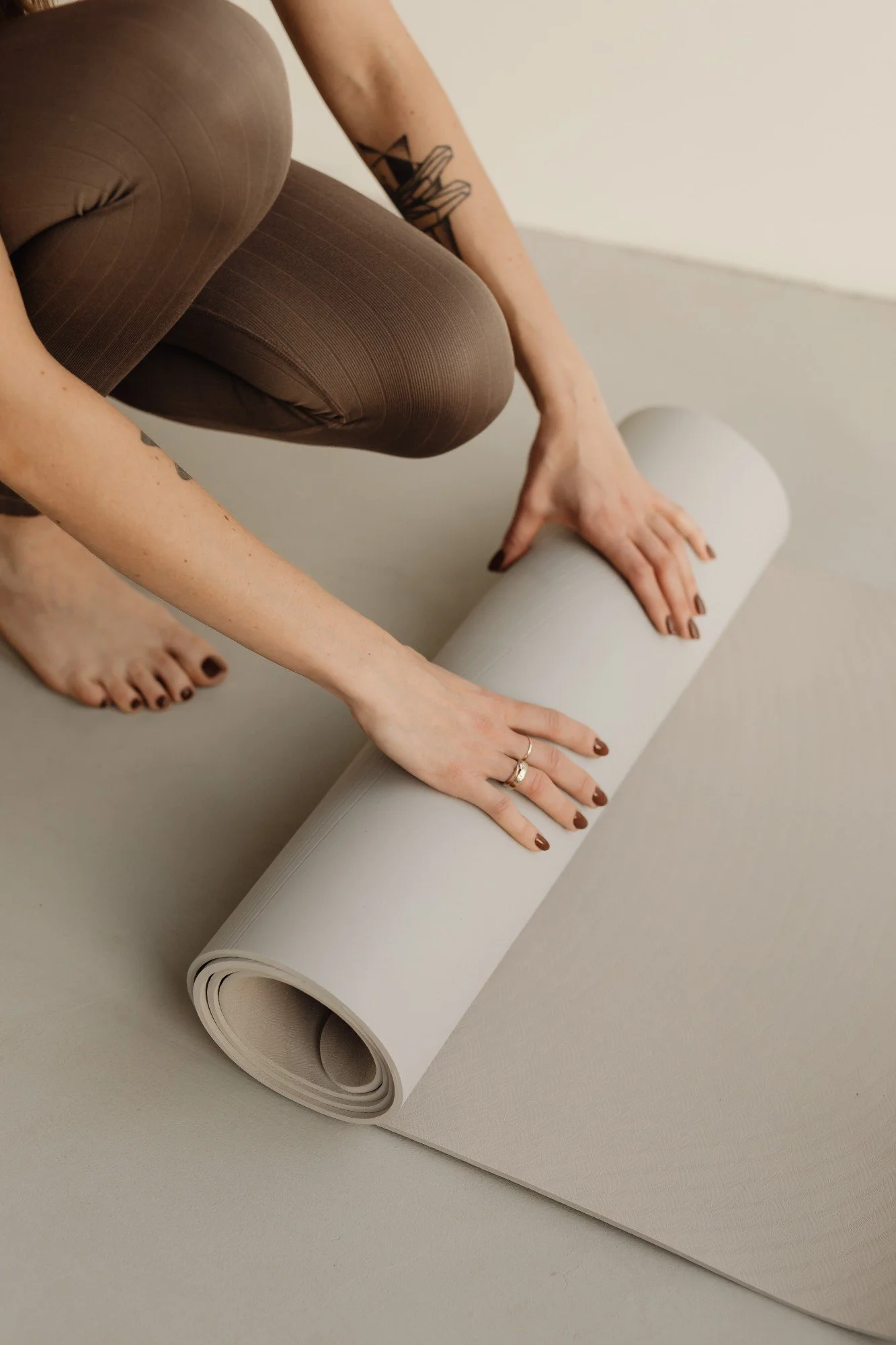 A person in brown leggings is unrolling a beige yoga mat on a gray surface. The person's hands are visible, with rings on their fingers. The person's tattoos are visible on their right arm.