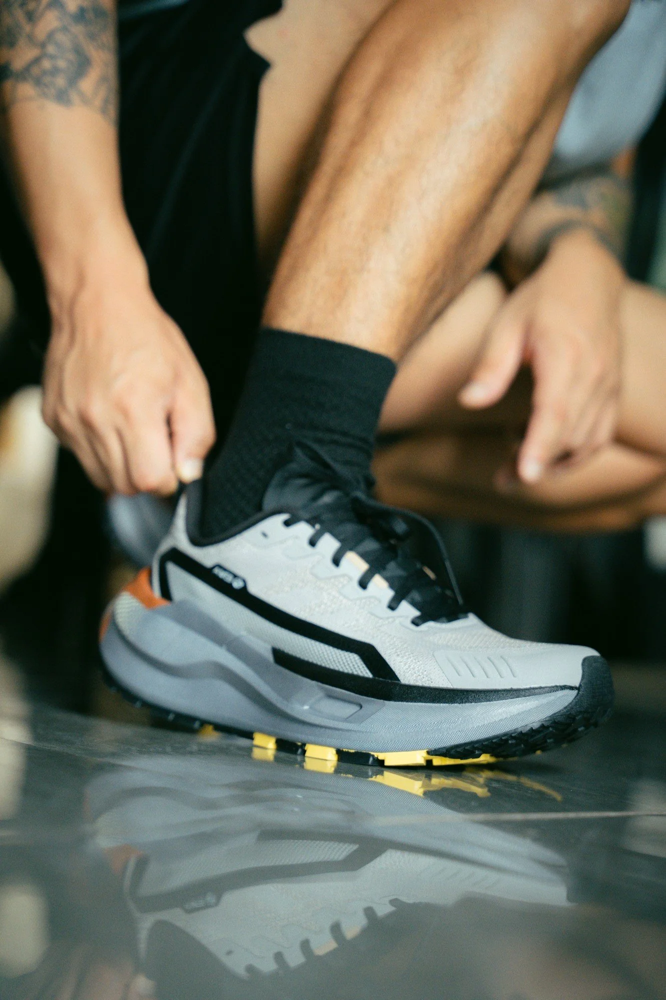 A person is tying their shoelaces on a white athletic shoe with black, gray, and orange accents. The person is wearing black socks and has tattoos on their arm. The setting appears to be indoors, possibly at a gym or sports facility.