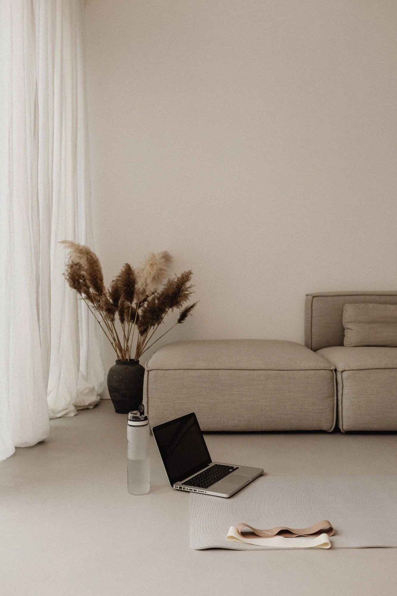 A minimalist living room with a beige sofa, a black vase of dried pampas grass, a laptop, a white water bottle, and rolled-up yoga mat on a light-colored rug, with sheer white curtains and a cream-colored wall in the background.