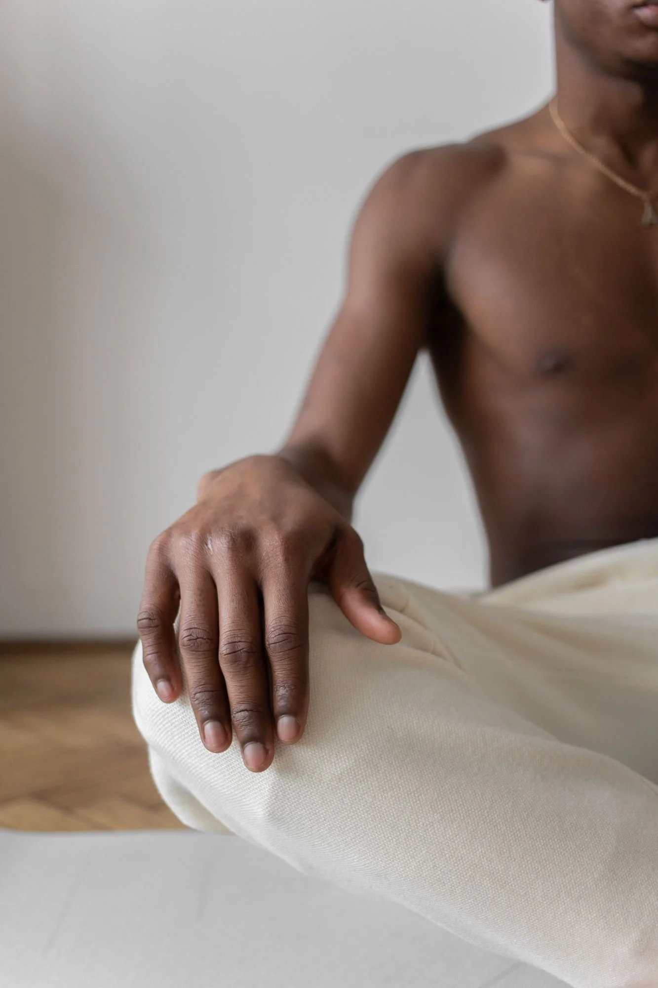 Close-up of a person doing yoga, with one hand resting on their knee and the other hand out of the frame. The individual has dark skin and is wearing a gold necklace and beige pants, against a neutral background.