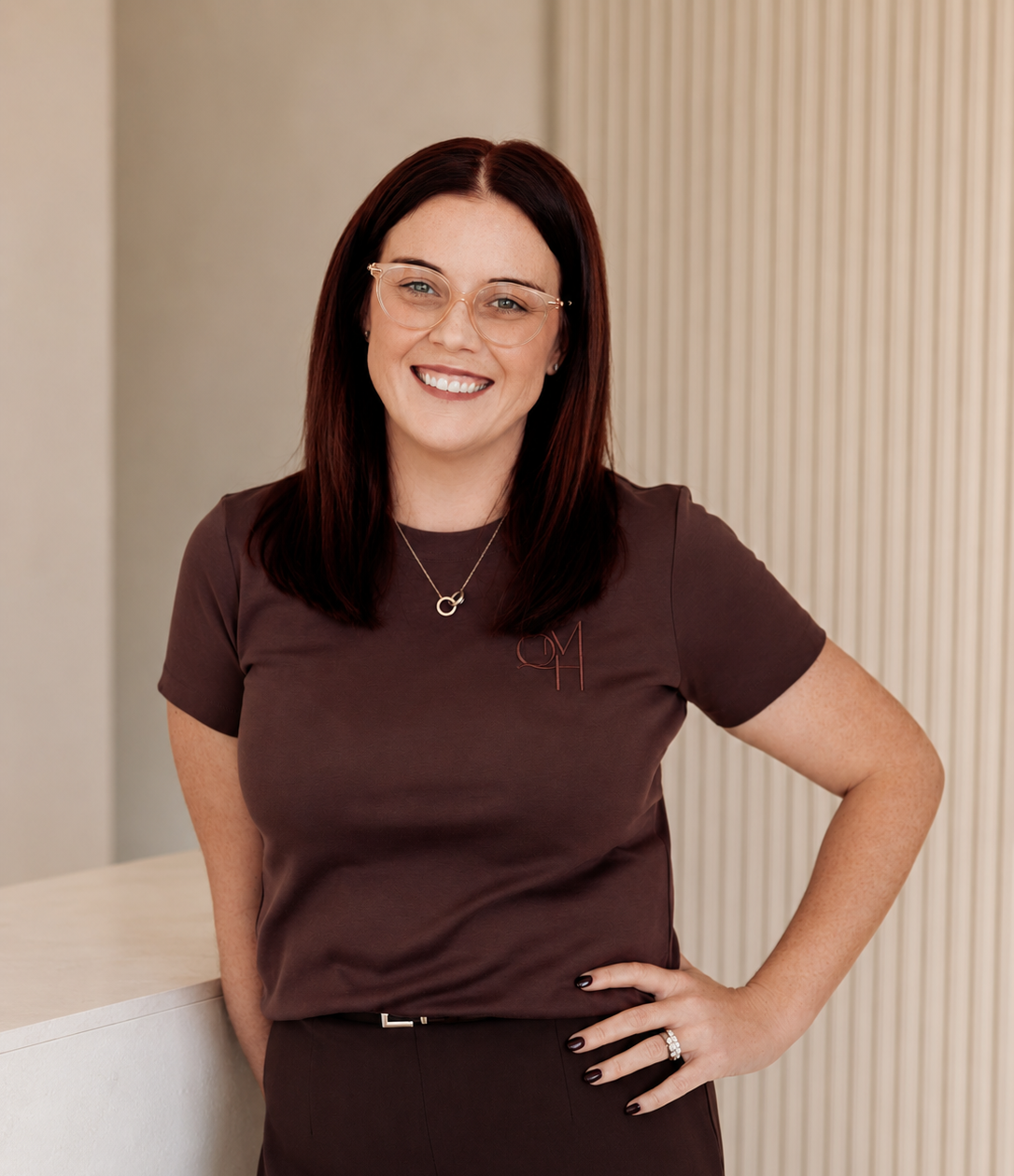 A smiling woman with brown hair, wearing glasses, a brown shirt, and a necklace, standing with her hand on her hip in an indoor setting.