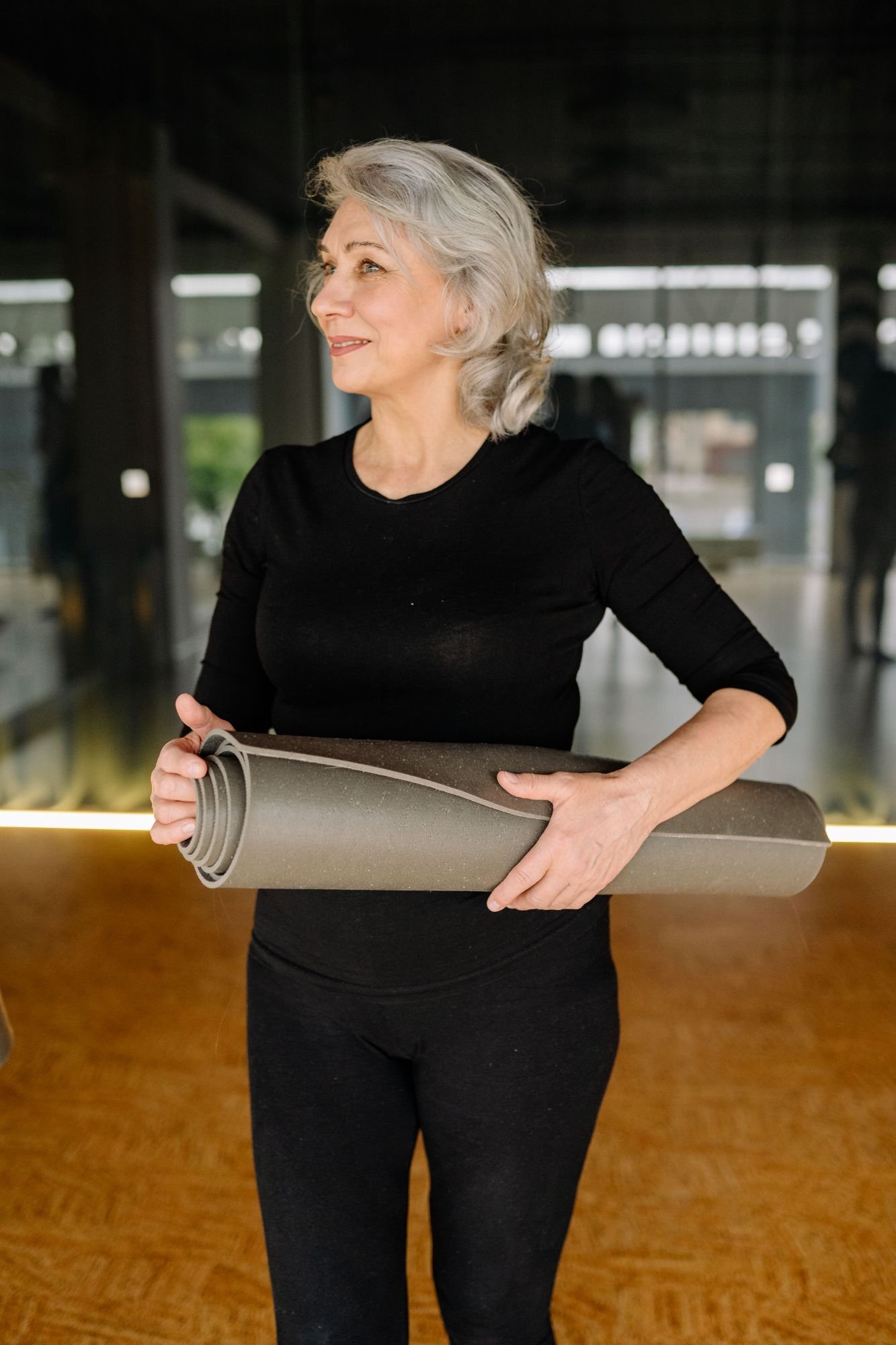 An older woman with gray hair smiling and holding a rolled gray yoga mat, standing indoors.