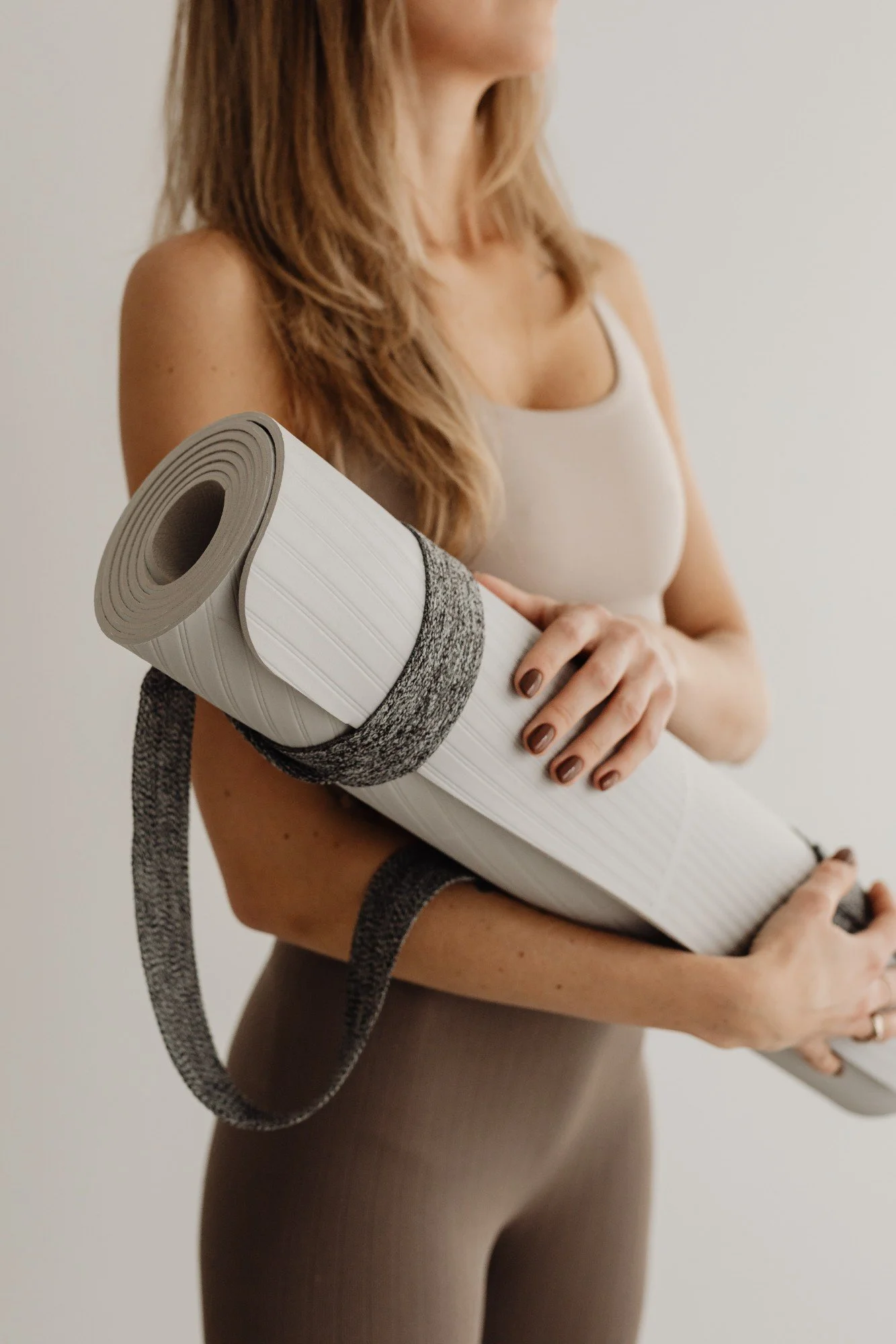A woman with long hair holding a rolled-up yoga mat, wearing a tank top and leggings.